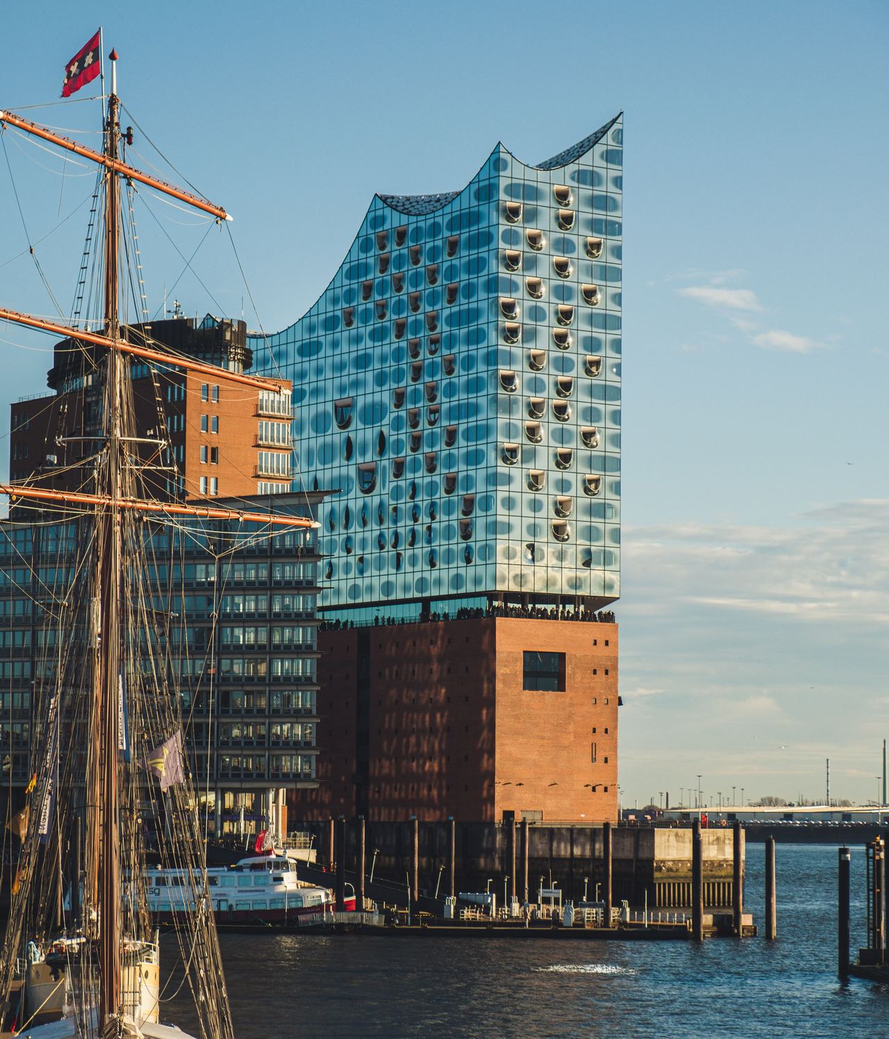 Modernes Gebäude mit wellenförmiger Glasfassade am Wasser, vor einem klaren Himmel, neben Segelschiffen im Hafen. Modernes Gebäude mit wellenförmiger Glasfassade am Wasser, vor einem klaren Himmel, neben Segelschiffen im Hafen.