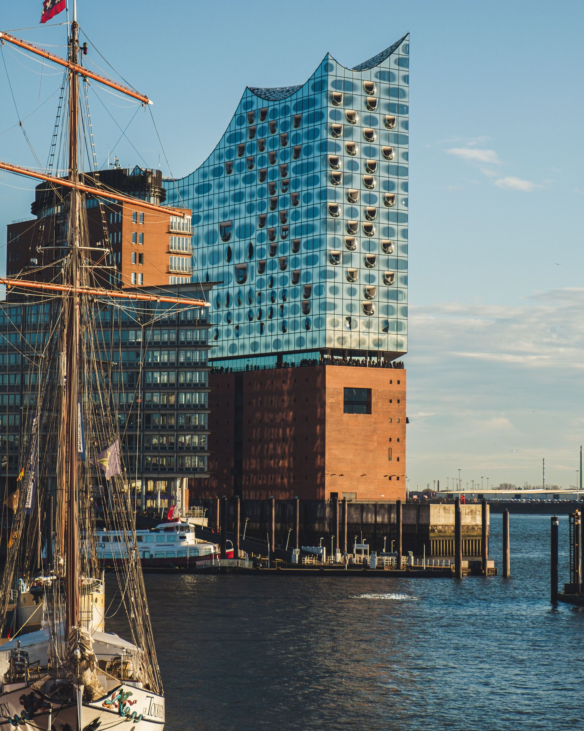 Modernes Gebäude mit wellenförmiger Glasfassade am Wasser, vor einem klaren Himmel, neben Segelschiffen im Hafen.