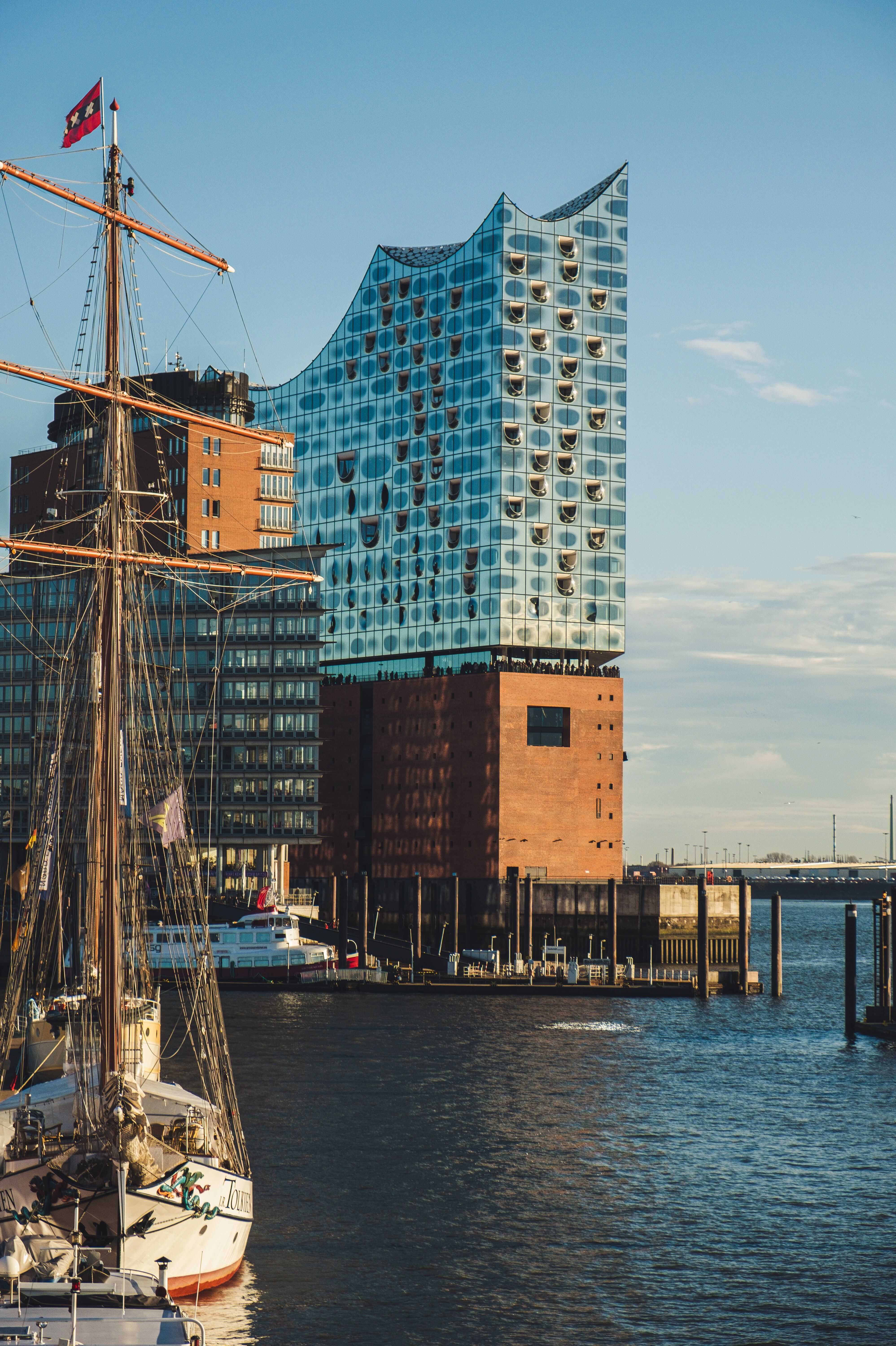 Modernes Gebäude mit wellenförmiger Glasfassade am Wasser, vor einem klaren Himmel, neben Segelschiffen im Hafen.