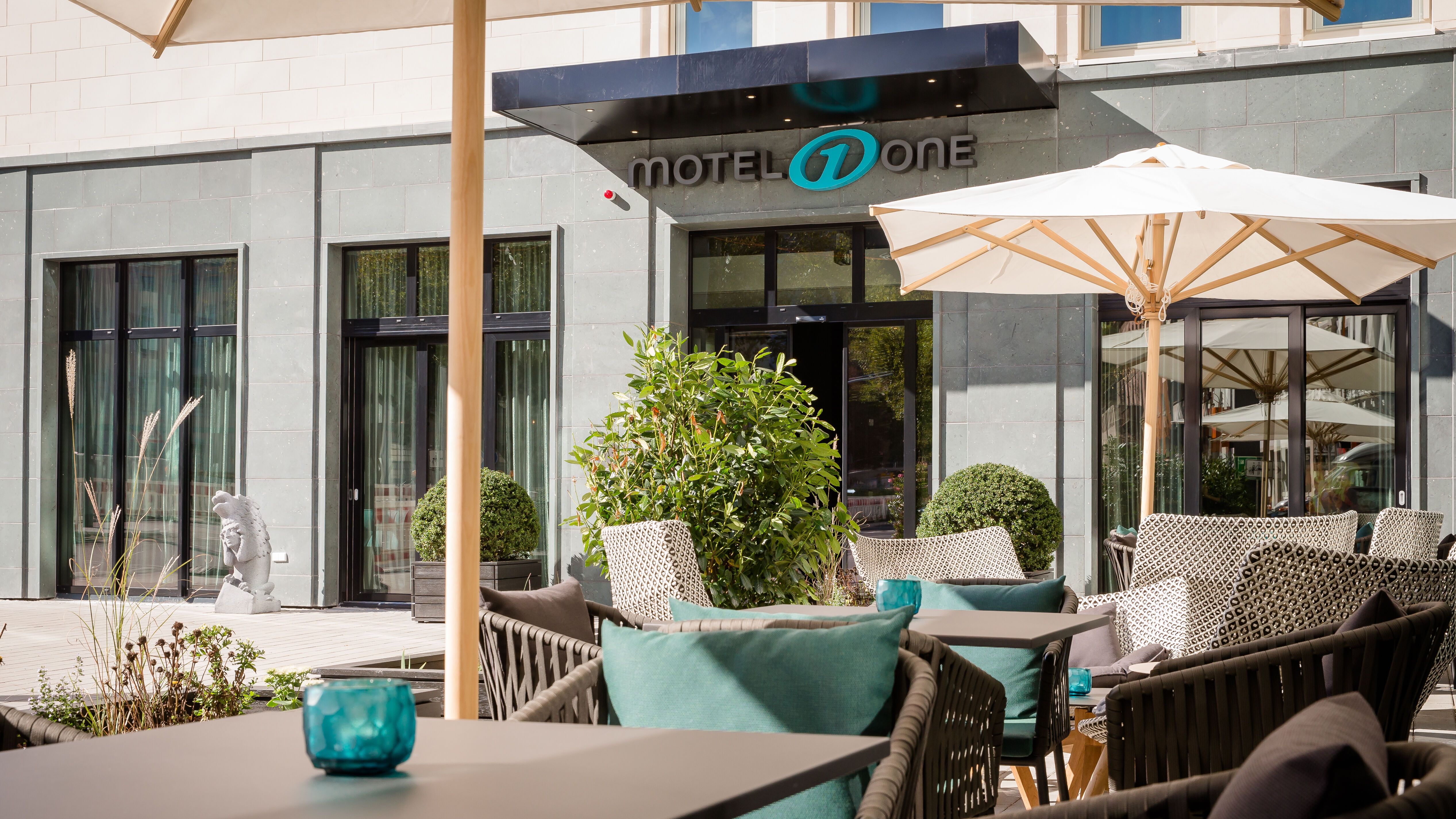 Außenbereich eines modernen Hotels mit gemütlichen Sitzgruppen, grünen Pflanzen und Sonnenschirmen, einladende Atmosphäre zum Entspannen vor dem Hoteleingang.