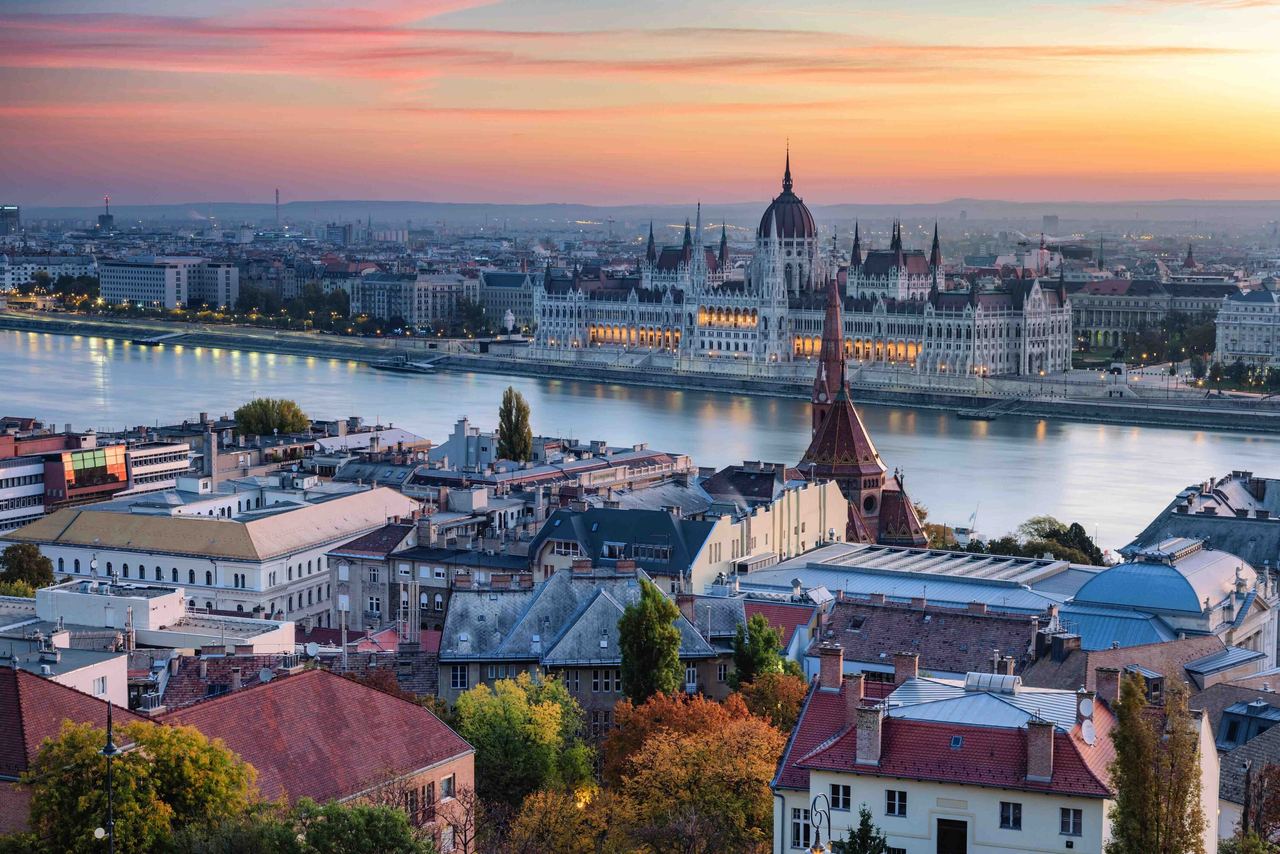 Panoramablick auf Budapest bei Sonnenuntergang mit dem beleuchteten ungarischen Parlament am Donauufer, umgeben von historischen Gebäuden und einem farbenfrohen Abendhimmel.
