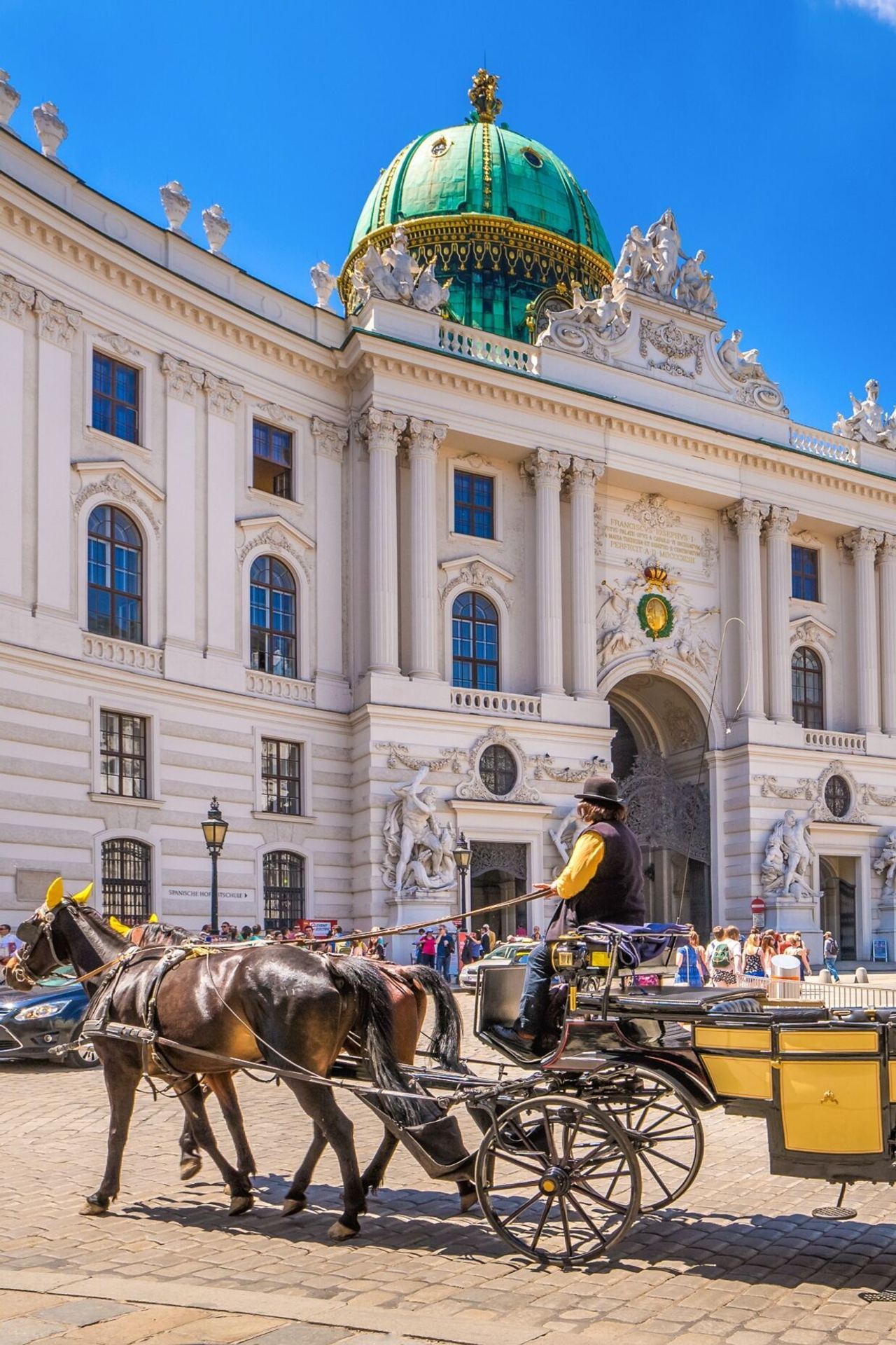 Vor dem historischen Hofburg-Palast mit grünen Kuppeln steht eine traditionelle Fiaker-Kutsche mit zwei Pferden, umgeben von Touristen bei strahlendem Himmel.