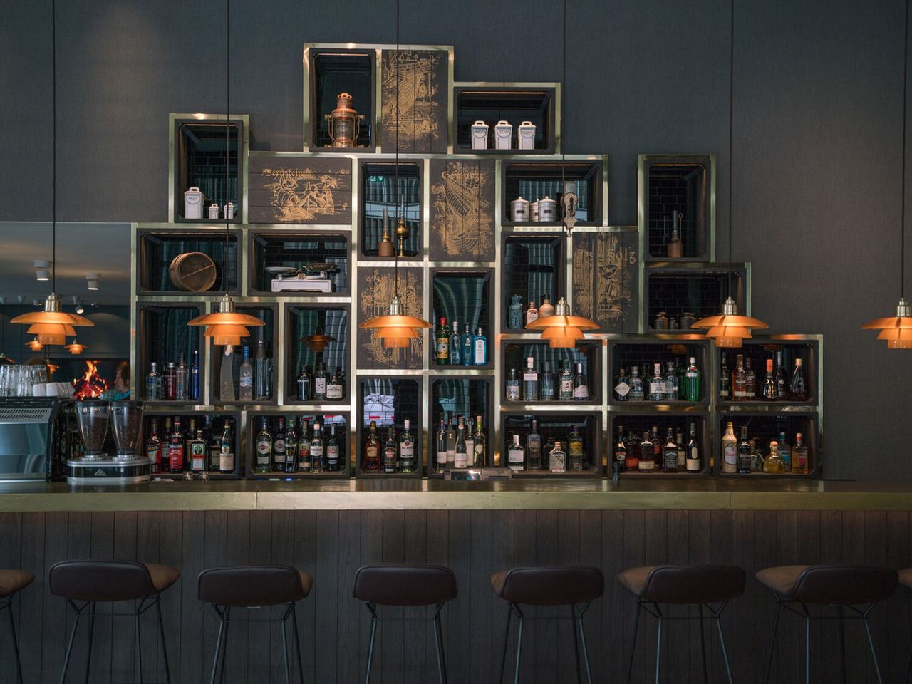 Motel One Hotelbar mit stilvoll angeordneten Regalen voller Spirituosen und edlen Flaschen, warm beleuchtet durch orangefarbene Hängeleuchten, stilvolle Barhocker davor.