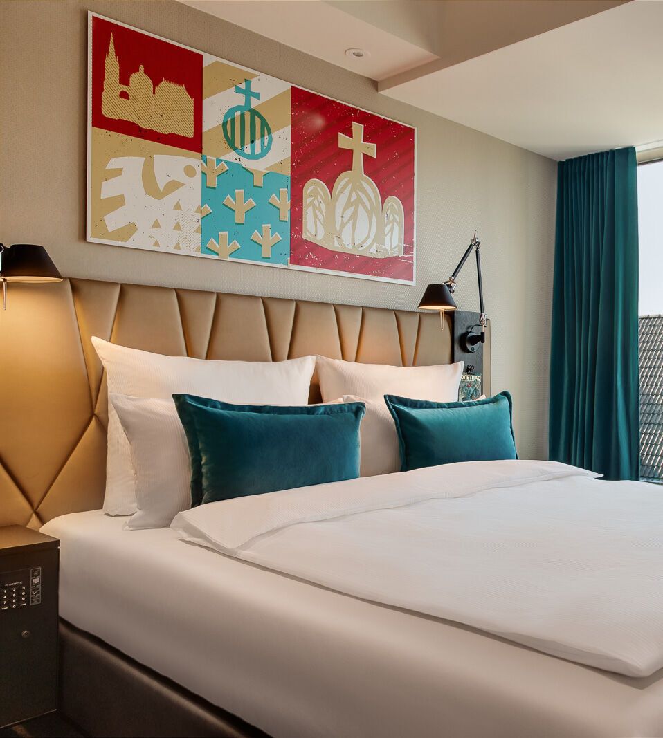 Helles Motel One Hotelzimmer mit großem, gemütlich bezogenem Doppelbett, moderner Wanddekoration und bodentiefen Fenstern mit Blick auf historische Kirchturmspitzen.