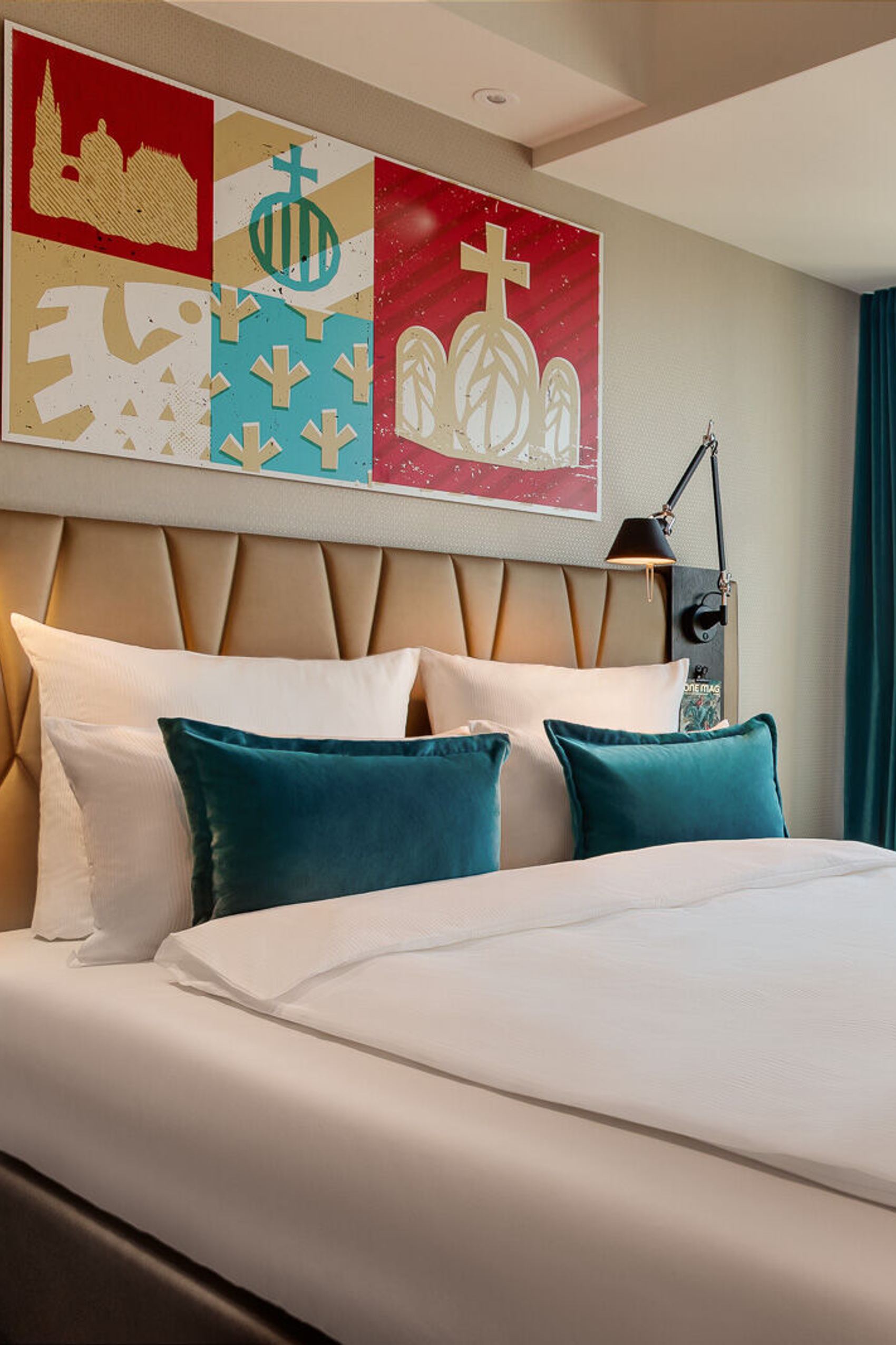 Helles Motel One Hotelzimmer mit großem, gemütlich bezogenem Doppelbett, moderner Wanddekoration und bodentiefen Fenstern mit Blick auf historische Kirchturmspitzen.