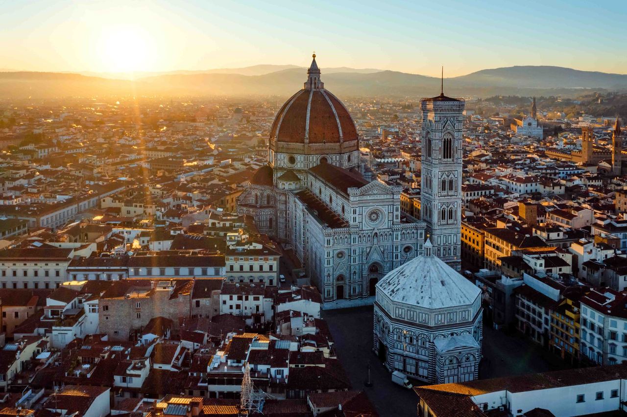 Luftaufnahme von Florenz bei Sonnenuntergang mit der markanten Kuppel der Kathedrale Santa Maria del Fiore, dem Baptisterium und dem Campanile, eingebettet in die historische Stadtlandschaft vor den sanften Hügeln im Hintergrund.
