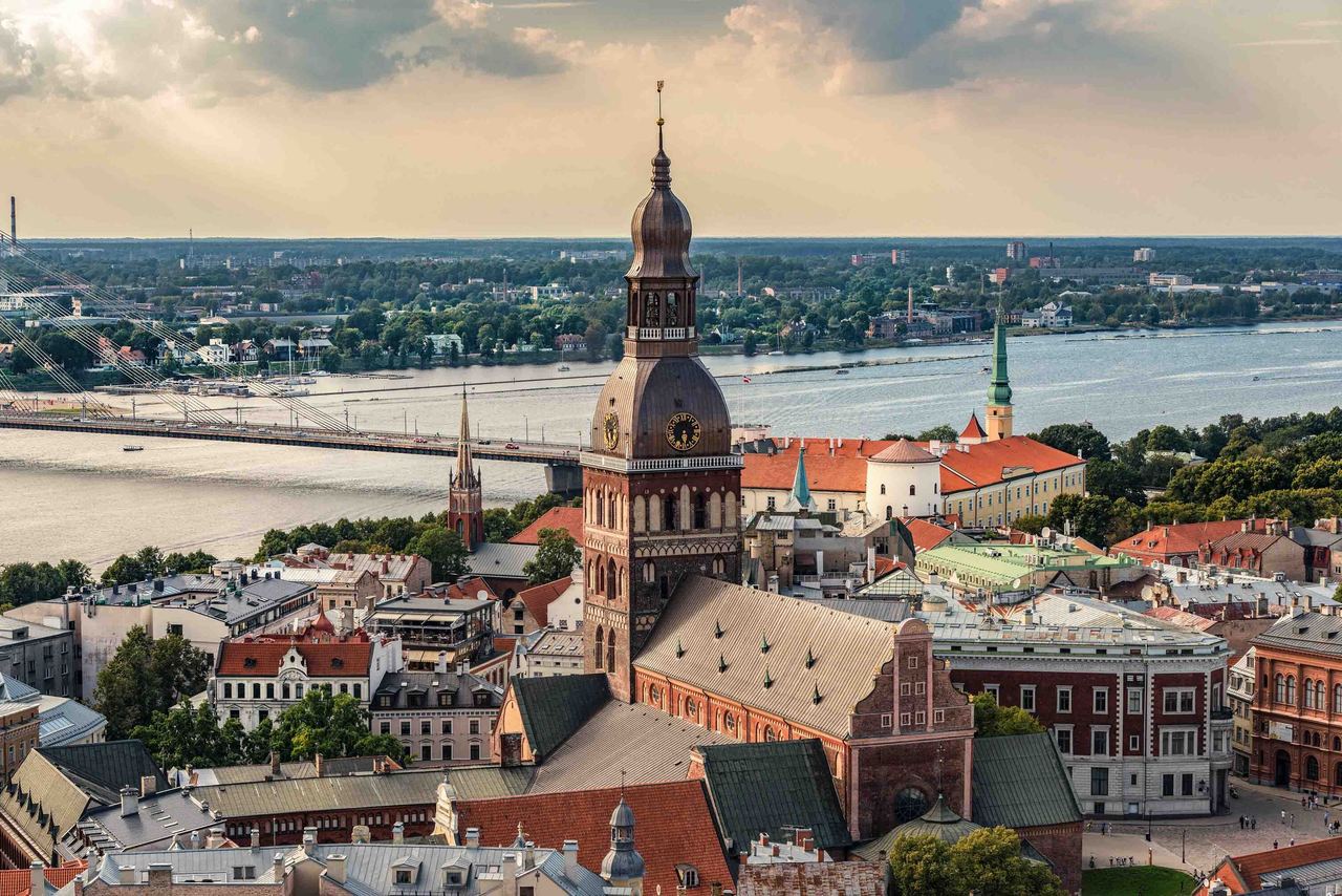 Blick über die Altstadt mit historischen Gebäuden und einem markanten Turm, im Hintergrund ein breiter Fluss und eine Brücke unter wolkigem Himmel.