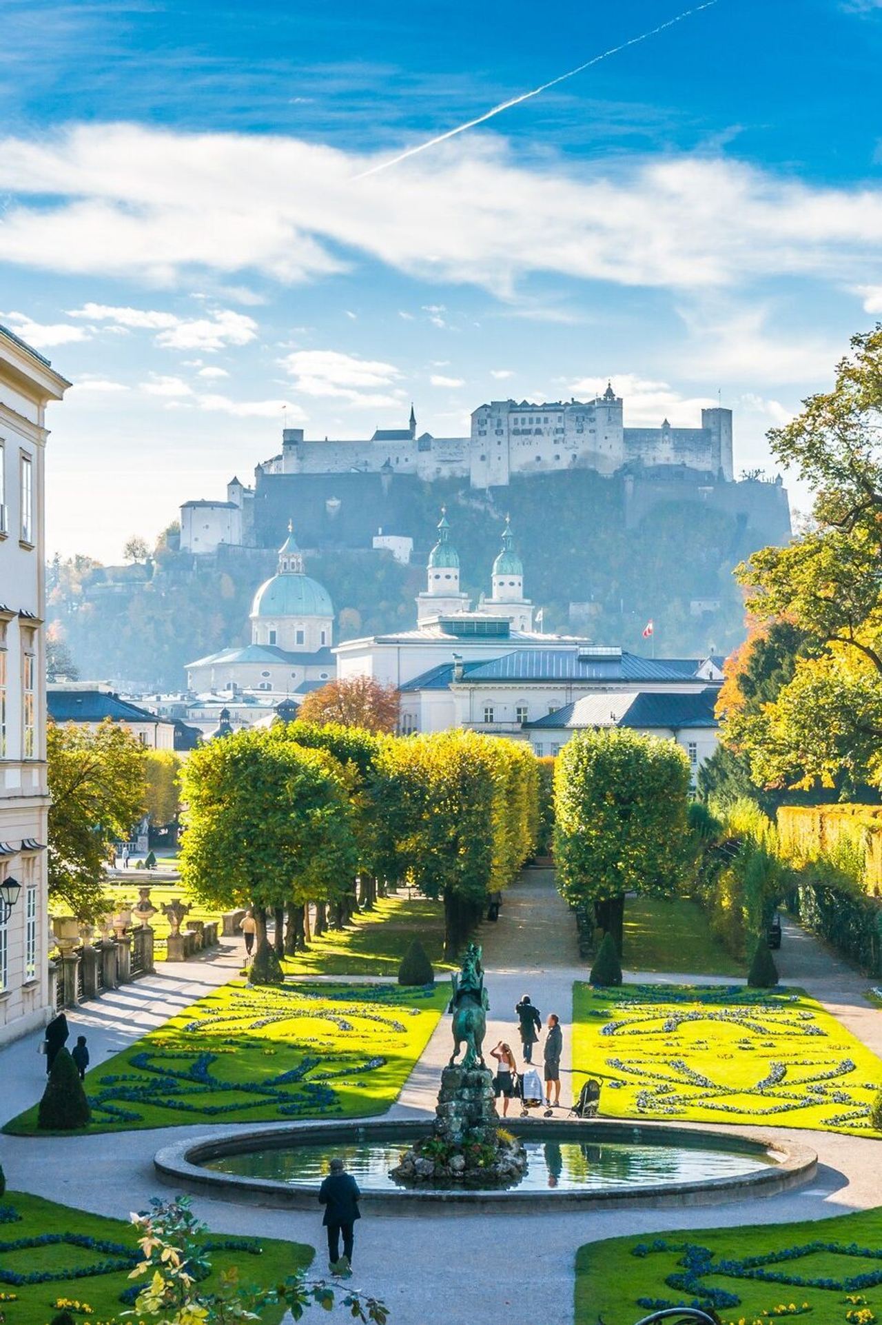 Blick auf einen gepflegten Barockgarten mit kunstvoll geschnittenen Hecken und Springbrunnen, umgeben von historischen Gebäuden, mit der Festung Hohensalzburg im Hintergrund unter blauem Himmel. Blick auf einen gepflegten Barockgarten mit kunstvoll geschnittenen Hecken und Springbrunnen, umgeben von historischen Gebäuden, mit der Festung Hohensalzburg im Hintergrund unter blauem Himmel.