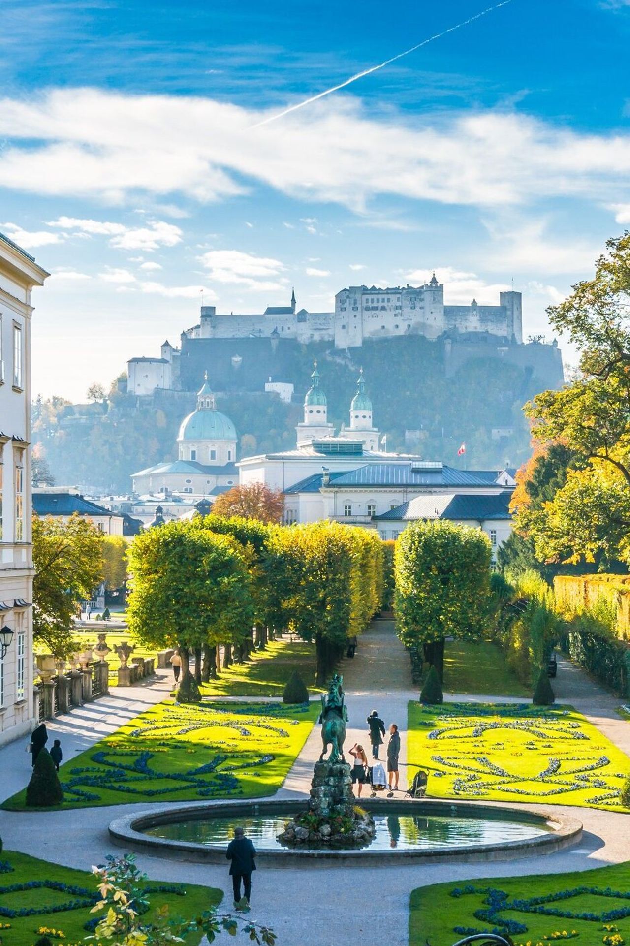 Blick auf einen gepflegten Barockgarten mit kunstvoll geschnittenen Hecken und Springbrunnen, umgeben von historischen Gebäuden, mit der Festung Hohensalzburg im Hintergrund unter blauem Himmel.