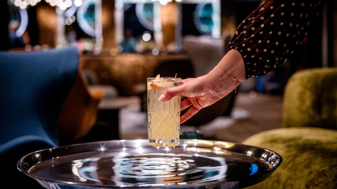Hand, die in stilvoll eingerichteter Lounge ein erfrischendes, dekoriertes Cocktailglas von einem glänzenden Silbertablett nimmt.