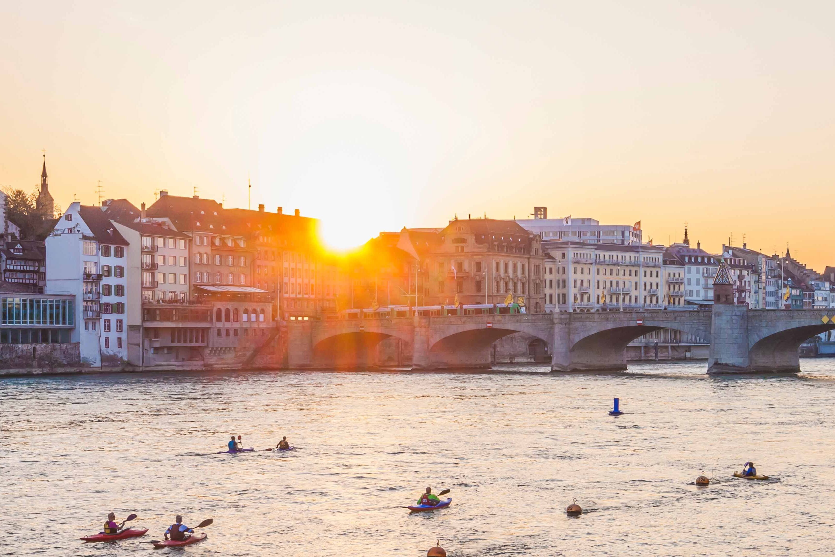 Sonnenuntergang über einem Fluss mit einer historischen Stadt im Hintergrund. Menschen paddeln im Kanu auf dem ruhigen Wasser, während eine Brücke die Ufer verbindet. 