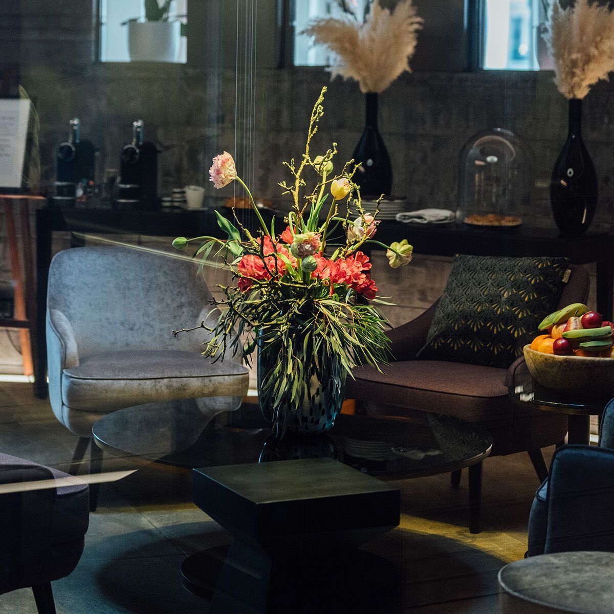 Gemütliche Lounge mit moderner Beleuchtung, Sofa, Sesseln und einem Sofa-Tisch mit farbenfrohem Blumenstrauß. Ruhige Atmosphäre, ideal zum Entspannen oder für Gespräche.