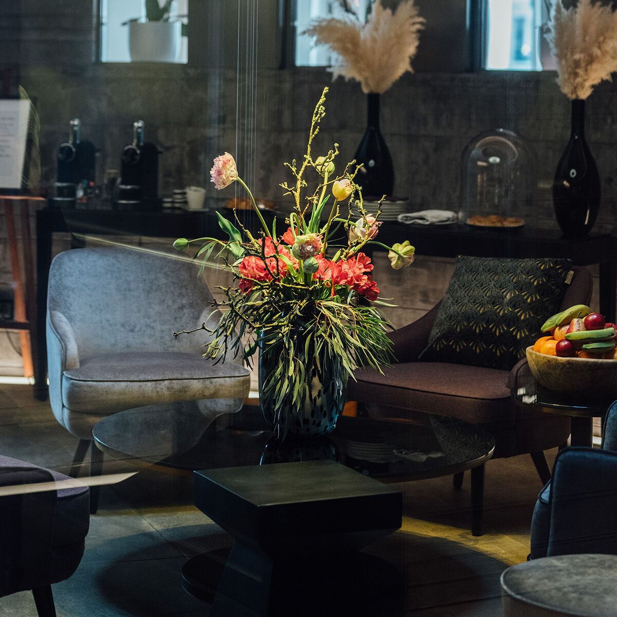 Gemütliche Lounge mit moderner Beleuchtung, Sofa, Sesseln und einem Sofa-Tisch mit farbenfrohem Blumenstrauß. Ruhige Atmosphäre, ideal zum Entspannen oder für Gespräche.