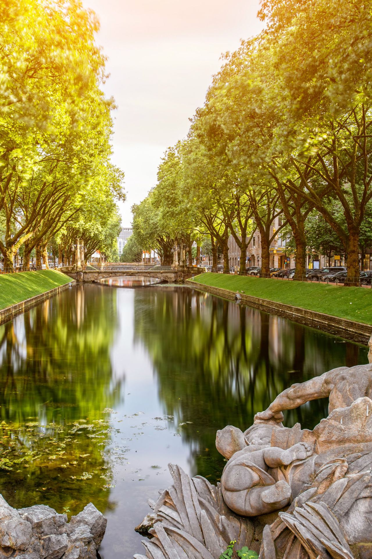 Fluss mit ruhigem Wasser, umgeben von grünen Bäumen an beiden Ufern, davor eine kunstvolle Steinfigur, im Hintergrund moderne Gebäude und eine Fußgängerbrücke, alles in warmem Sonnenlicht.