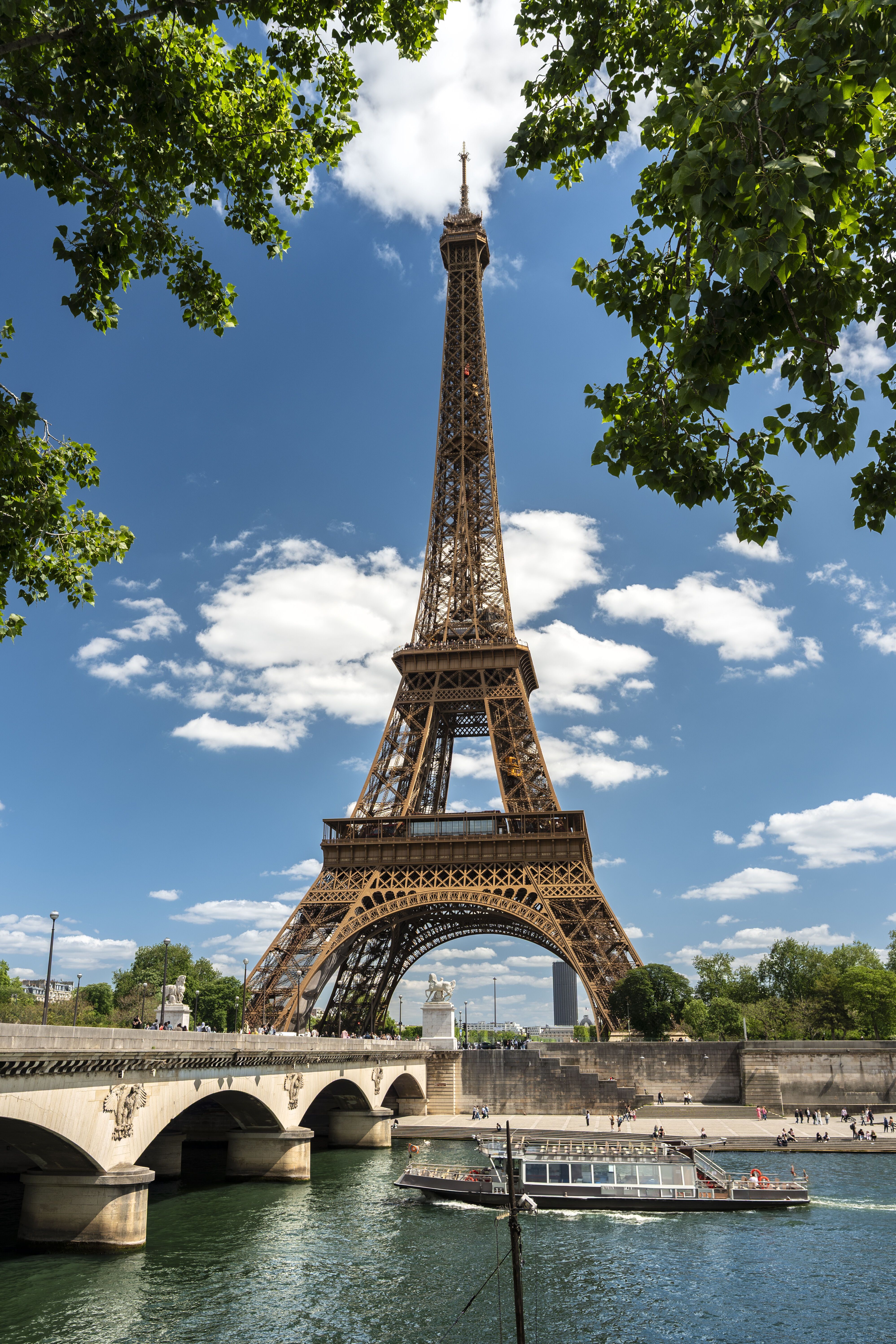 Der Eiffelturm ragt bei klarem Himmel über der Seine empor, umrahmt von grünen Baumzweigen. Eine Brücke überspannt den Fluss, auf dem ein Ausflugsschiff vorbeifährt.