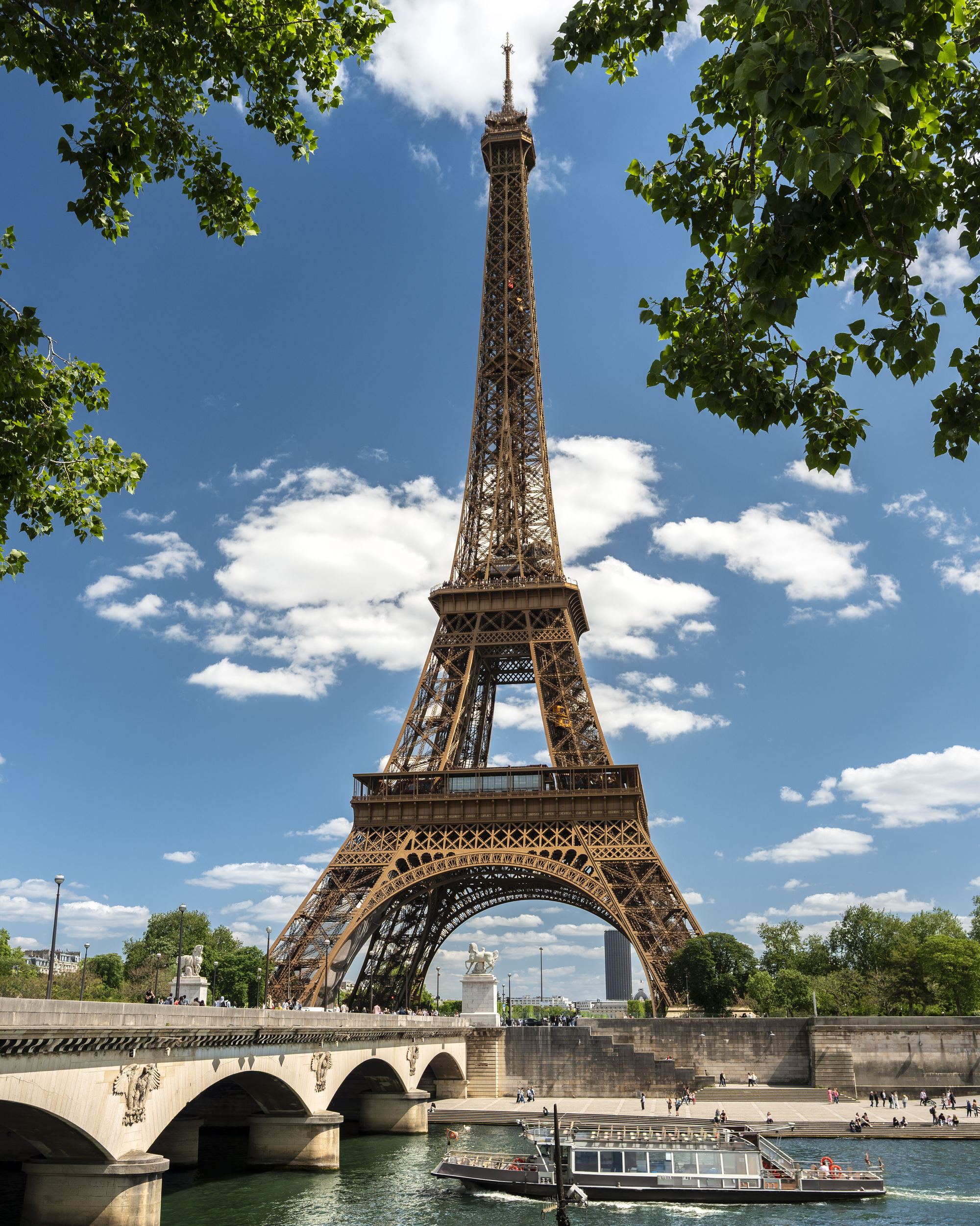 Der Eiffelturm ragt bei klarem Himmel über der Seine empor, umrahmt von grünen Baumzweigen. Eine Brücke überspannt den Fluss, auf dem ein Ausflugsschiff vorbeifährt.