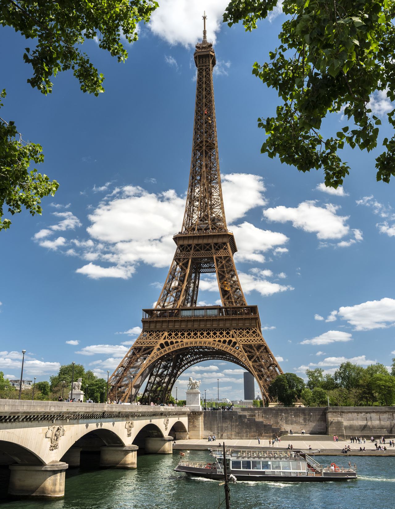 Der Eiffelturm ragt bei klarem Himmel über der Seine empor, umrahmt von grünen Baumzweigen. Eine Brücke überspannt den Fluss, auf dem ein Ausflugsschiff vorbeifährt. Der Eiffelturm ragt bei klarem Himmel über der Seine empor, umrahmt von grünen Baumzweigen. Eine Brücke überspannt den Fluss, auf dem ein Ausflugsschiff vorbeifährt.