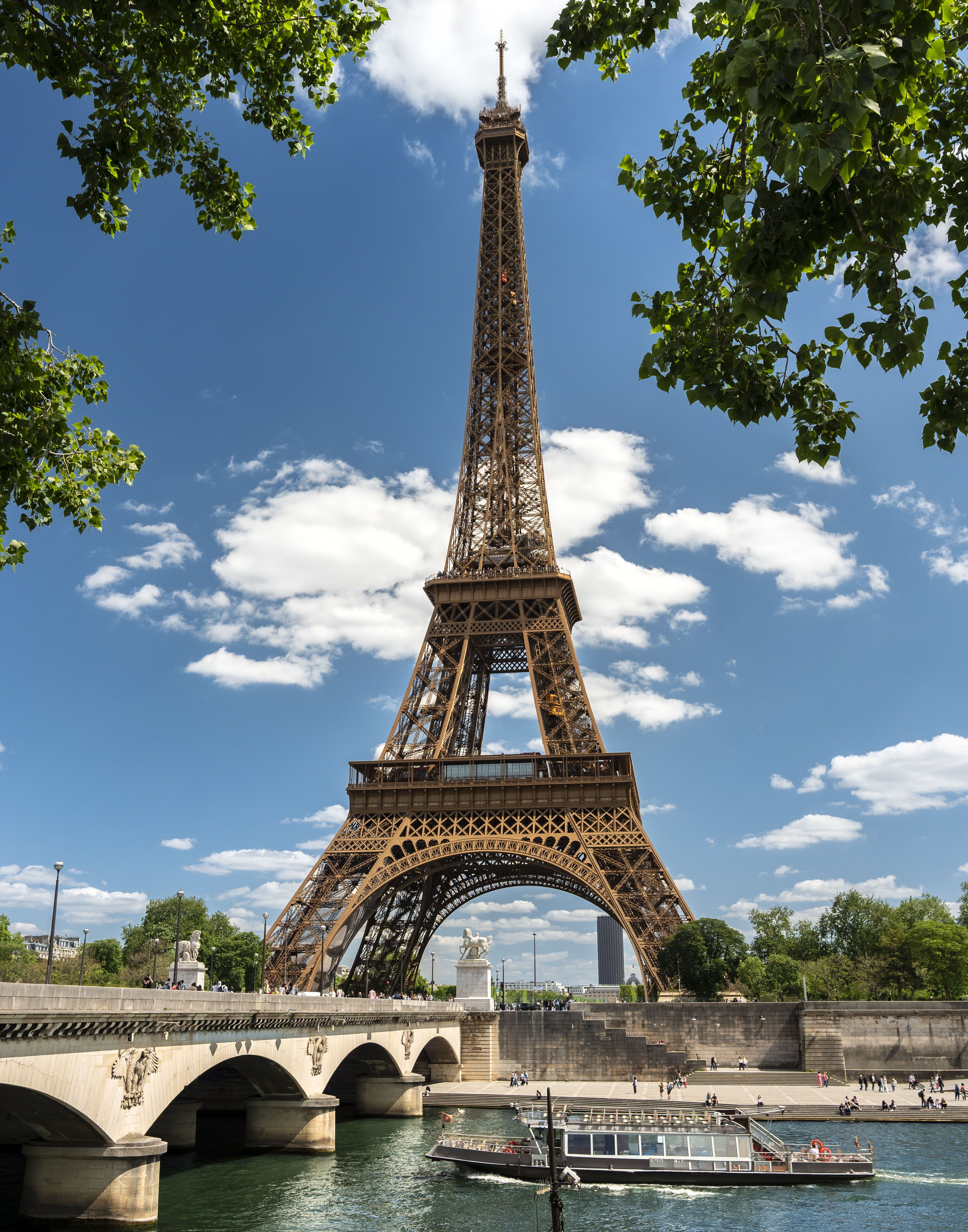Der Eiffelturm ragt bei klarem Himmel über der Seine empor, umrahmt von grünen Baumzweigen. Eine Brücke überspannt den Fluss, auf dem ein Ausflugsschiff vorbeifährt.