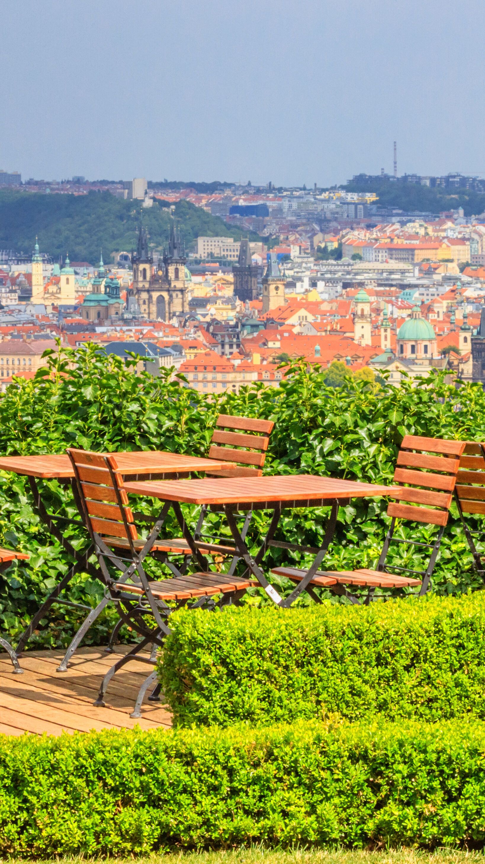 Dachterrasse mit Holztischen und Stühlen, Weinfass als Stehtisch, umgeben von grünen Hecken, mit weitem Blick über eine Stadtlandschaft im Hintergrund.