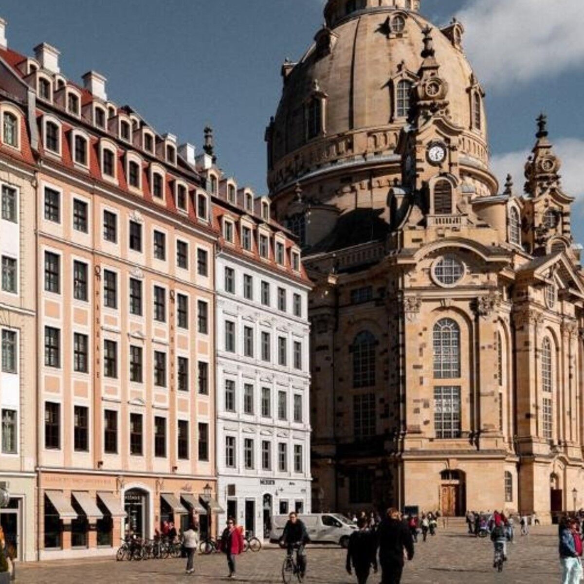 Historische Stadtsilhouette mit barocken Gebäuden und der imposanten Kuppelkirche im Hintergrund, belebter Platz mit Spaziergängern und Radfahrern bei klarem Himmel.