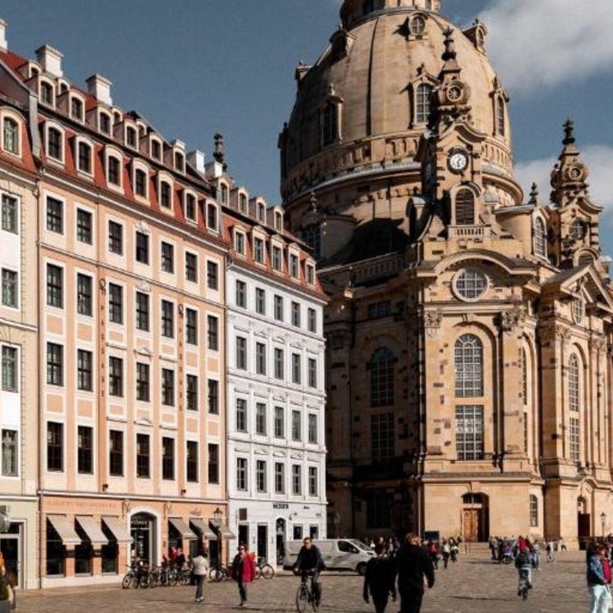 Historische Stadtsilhouette mit barocken Gebäuden und der imposanten Kuppelkirche im Hintergrund, belebter Platz mit Spaziergängern und Radfahrern bei klarem Himmel.