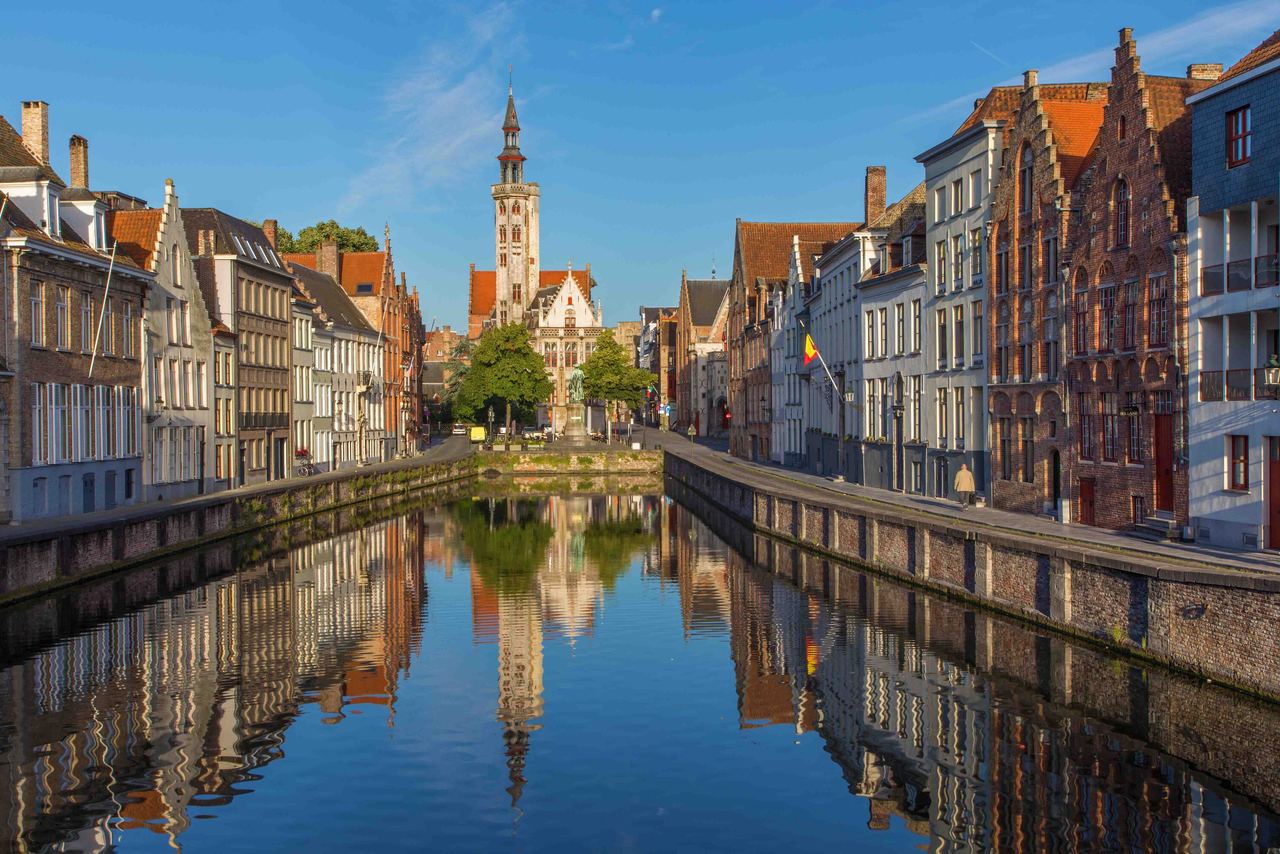Blick auf einen ruhigen Kanal in der Altstadt mit historischen Gebäuden und einem markanten Turm im Hintergrund, der sich im klaren Wasser spiegelt.