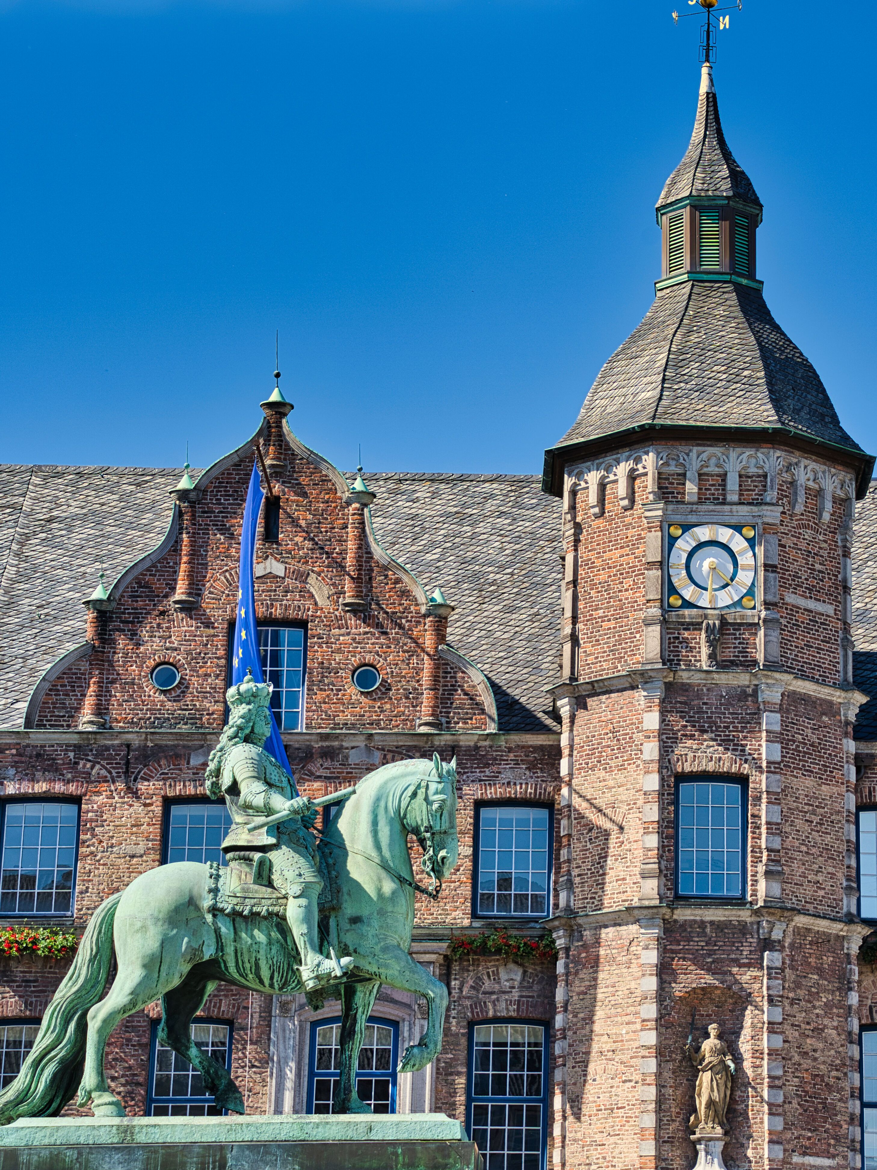 Historisches Backsteingebäude mit zentralem Turm und Uhr, davor eine Reiterstatue bei klarem, blauem Himmel.