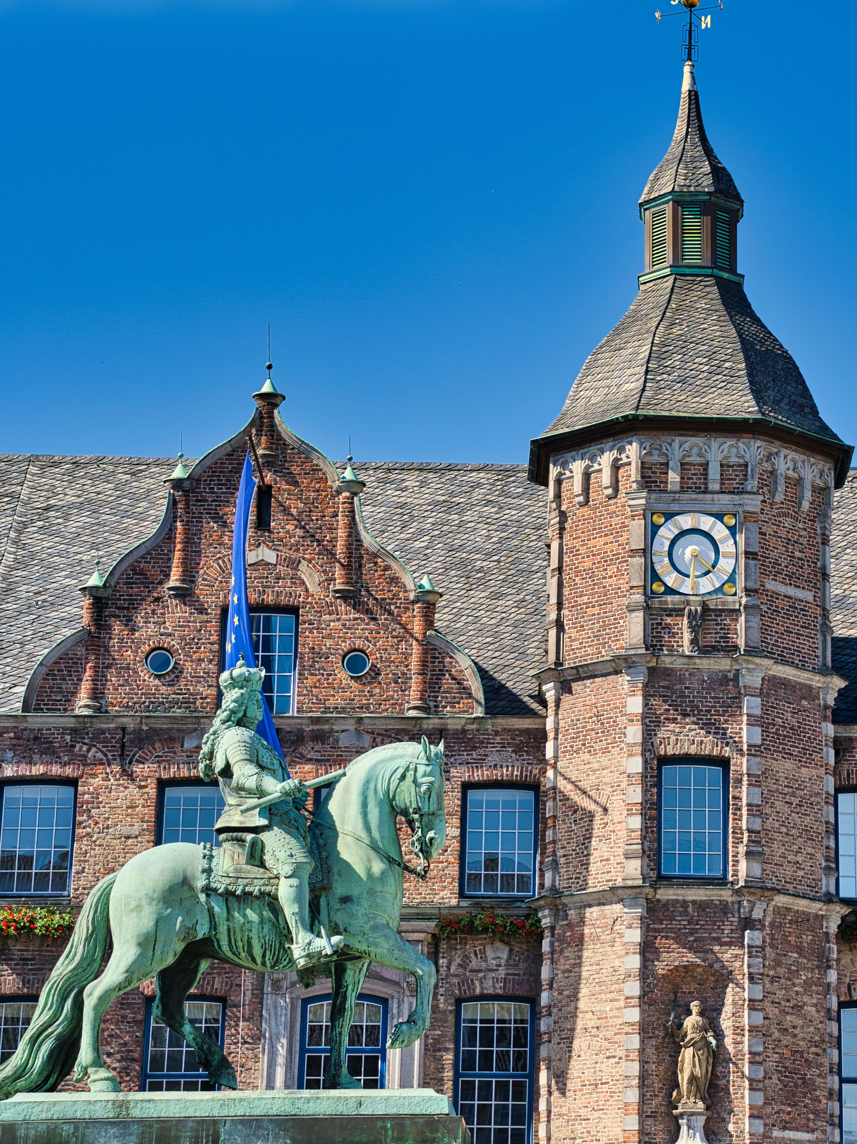 Historisches Backsteingebäude mit zentralem Turm und Uhr, davor eine Reiterstatue bei klarem, blauem Himmel.