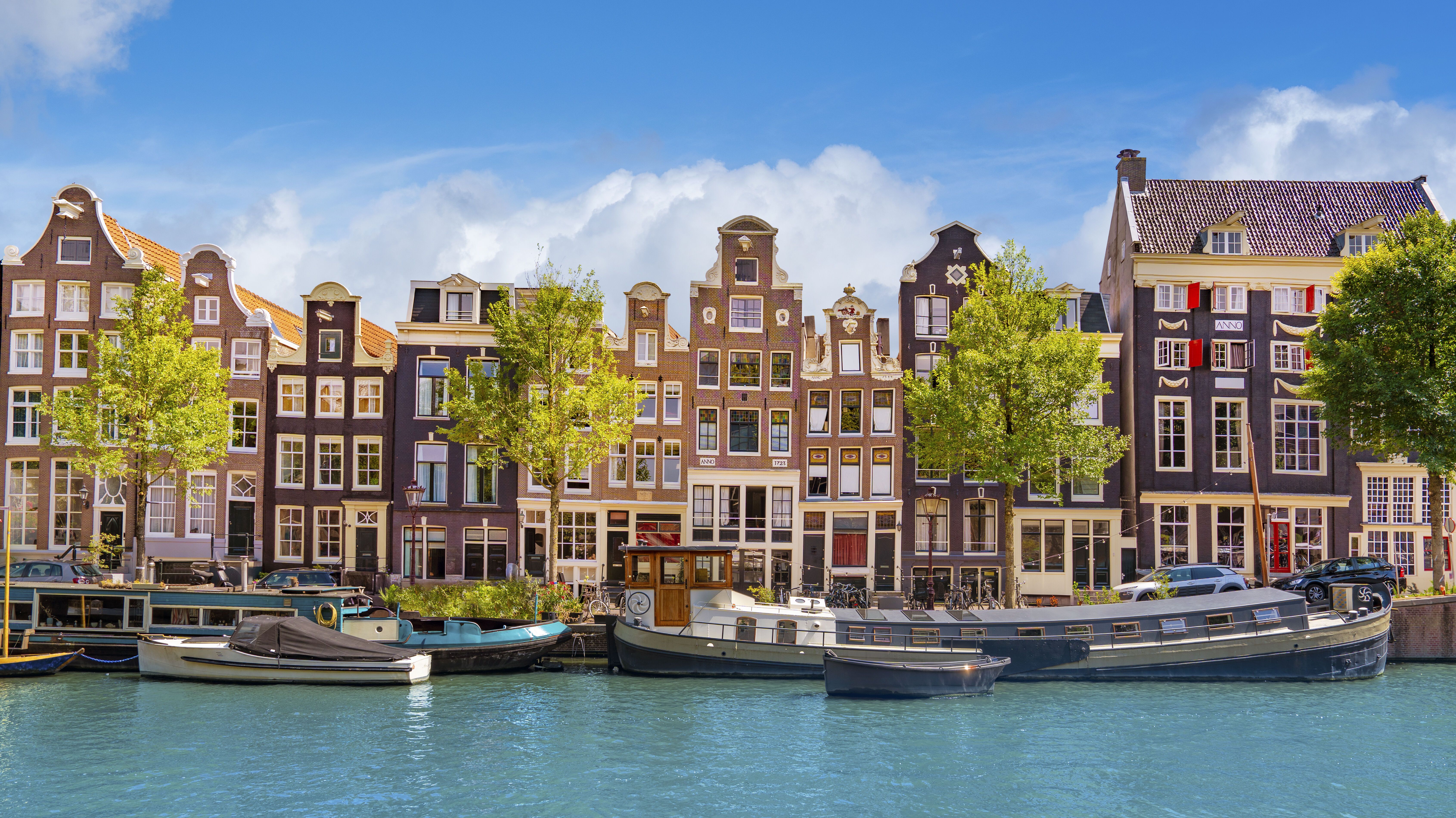 Bunte, historische Grachtenhäuser in Amsterdam mit markanten Giebeln entlang eines ruhigen Kanals, davor mehrere Boote im Wasser und grüne Bäume unter klarem, blauem Himmel.