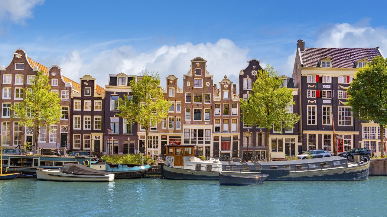 Bunte, historische Grachtenhäuser in Amsterdam mit markanten Giebeln entlang eines ruhigen Kanals, davor mehrere Boote im Wasser und grüne Bäume unter klarem, blauem Himmel.