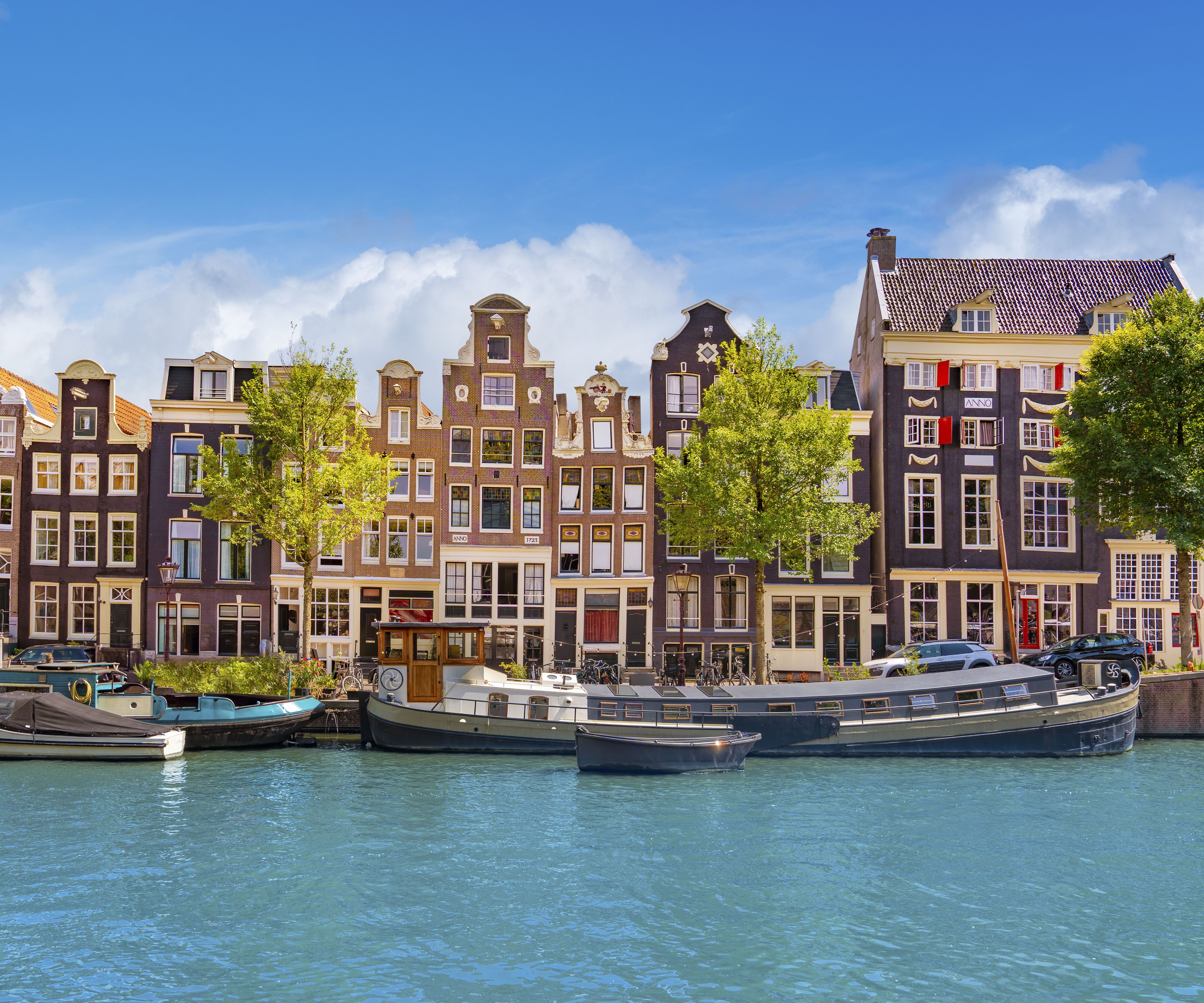 Bunte, historische Grachtenhäuser in Amsterdam mit markanten Giebeln entlang eines ruhigen Kanals, davor mehrere Boote im Wasser und grüne Bäume unter klarem, blauem Himmel.