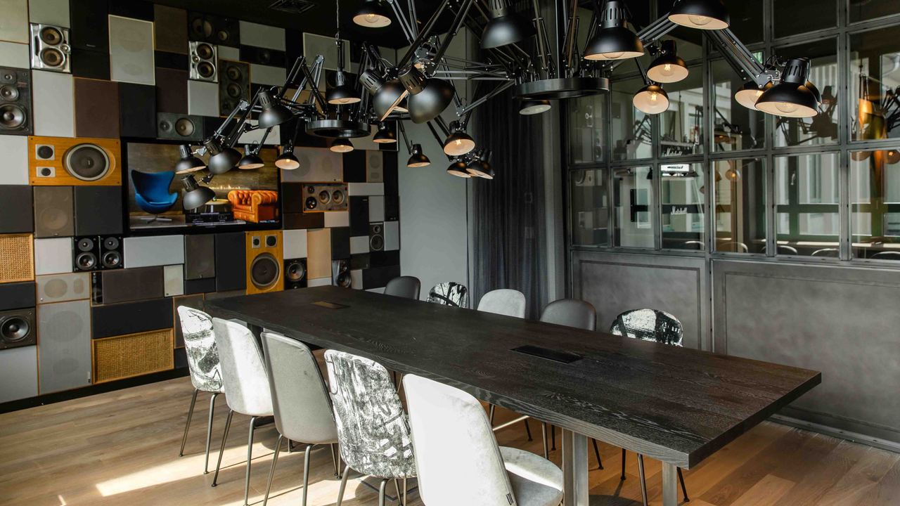 Motel One stilvoller Besprechungsraum mit großem dunklem Holztisch, acht verschieden gemusterte Stühle und kreativer Deckenbeleuchtung. Die Wand ist mit Lautsprechern und dekorativen Elementen gestaltet.