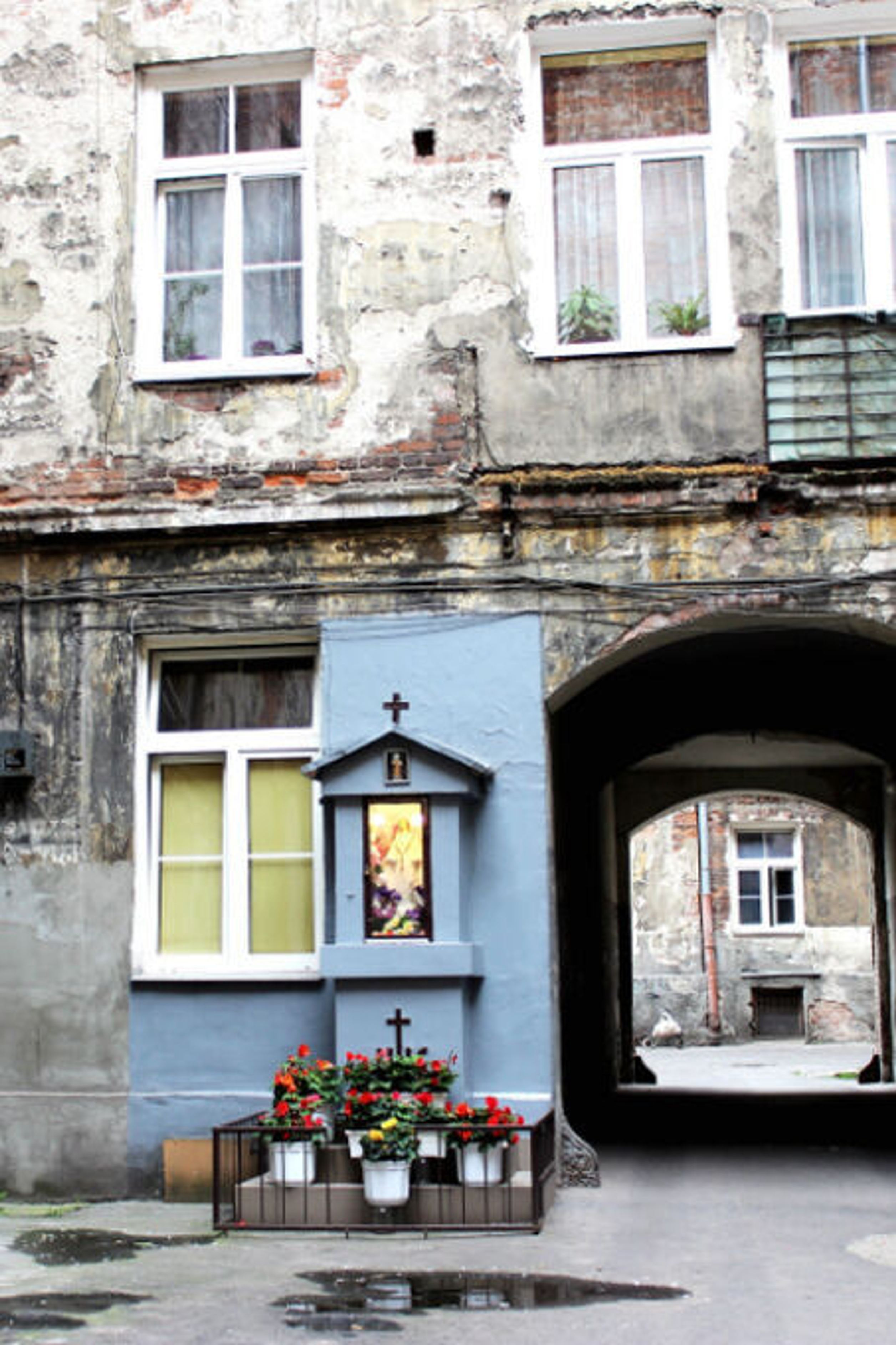 Innenhof eines alten, baufälligen Wohngebäudes mit verwitterten Fassaden. Ein kleines blau gestrichenes Wandaltar mit Blumen und Kerzen steht unter Fenstern, daneben ein dunkler Durchgang zum gegenüberliegenden Hof.