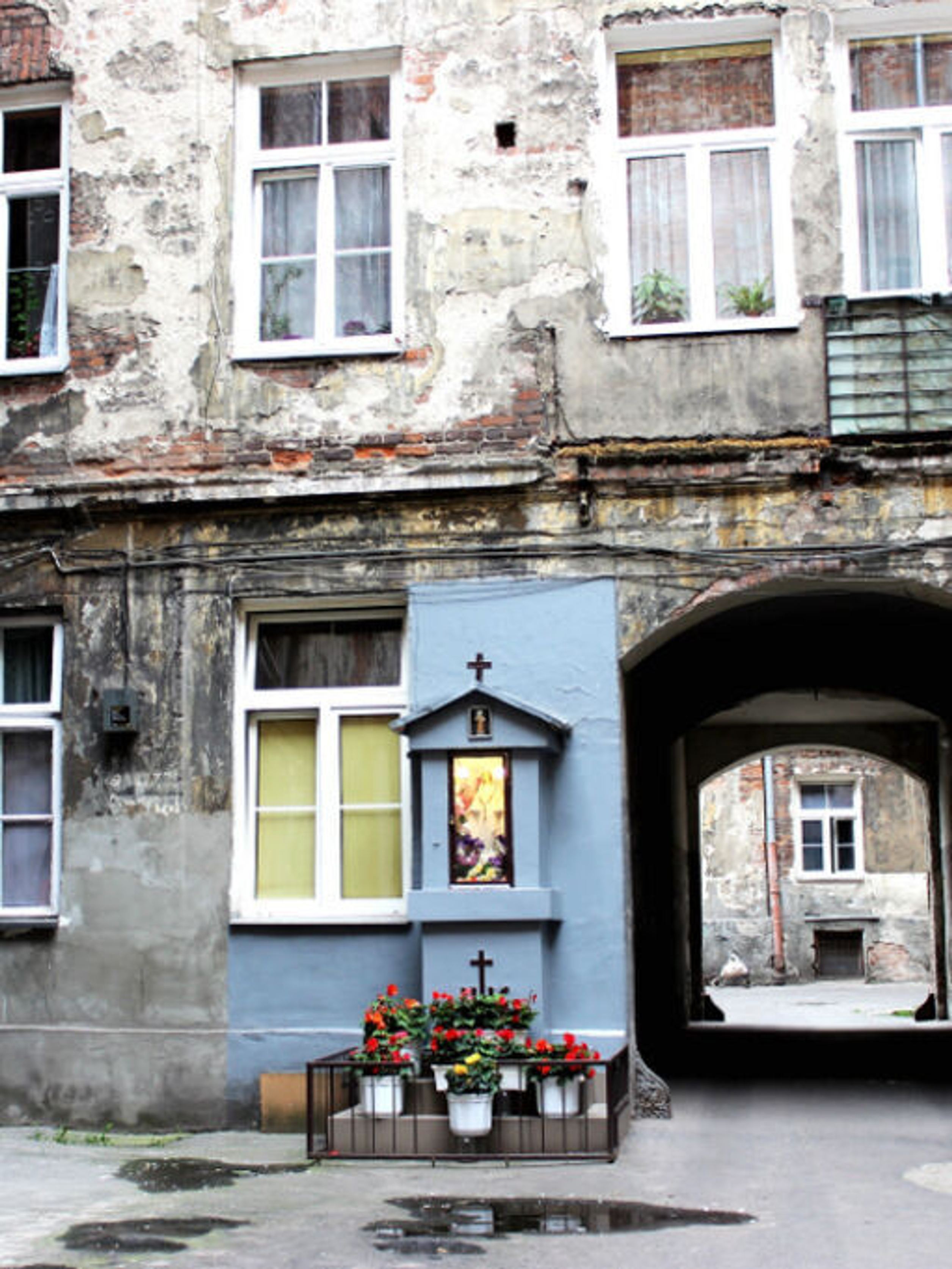 Innenhof eines alten, baufälligen Wohngebäudes mit verwitterten Fassaden. Ein kleines blau gestrichenes Wandaltar mit Blumen und Kerzen steht unter Fenstern, daneben ein dunkler Durchgang zum gegenüberliegenden Hof. Innenhof eines alten, baufälligen Wohngebäudes mit verwitterten Fassaden. Ein kleines blau gestrichenes Wandaltar mit Blumen und Kerzen steht unter Fenstern, daneben ein dunkler Durchgang zum gegenüberliegenden Hof.