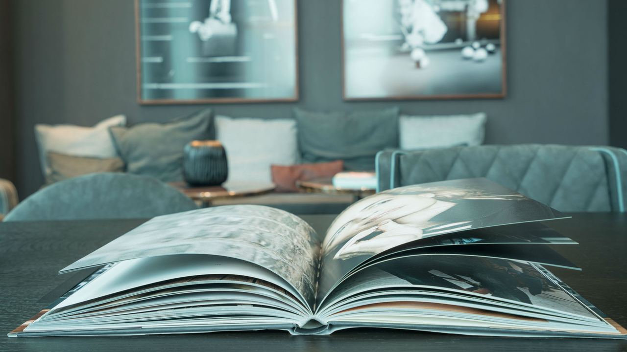 Aufgeschlagenes Fotobuch auf einem Tisch in einem gemütlichen eingerichteten Wohnzimmer mit Sofa und dekorativen Bildern an der Wand.