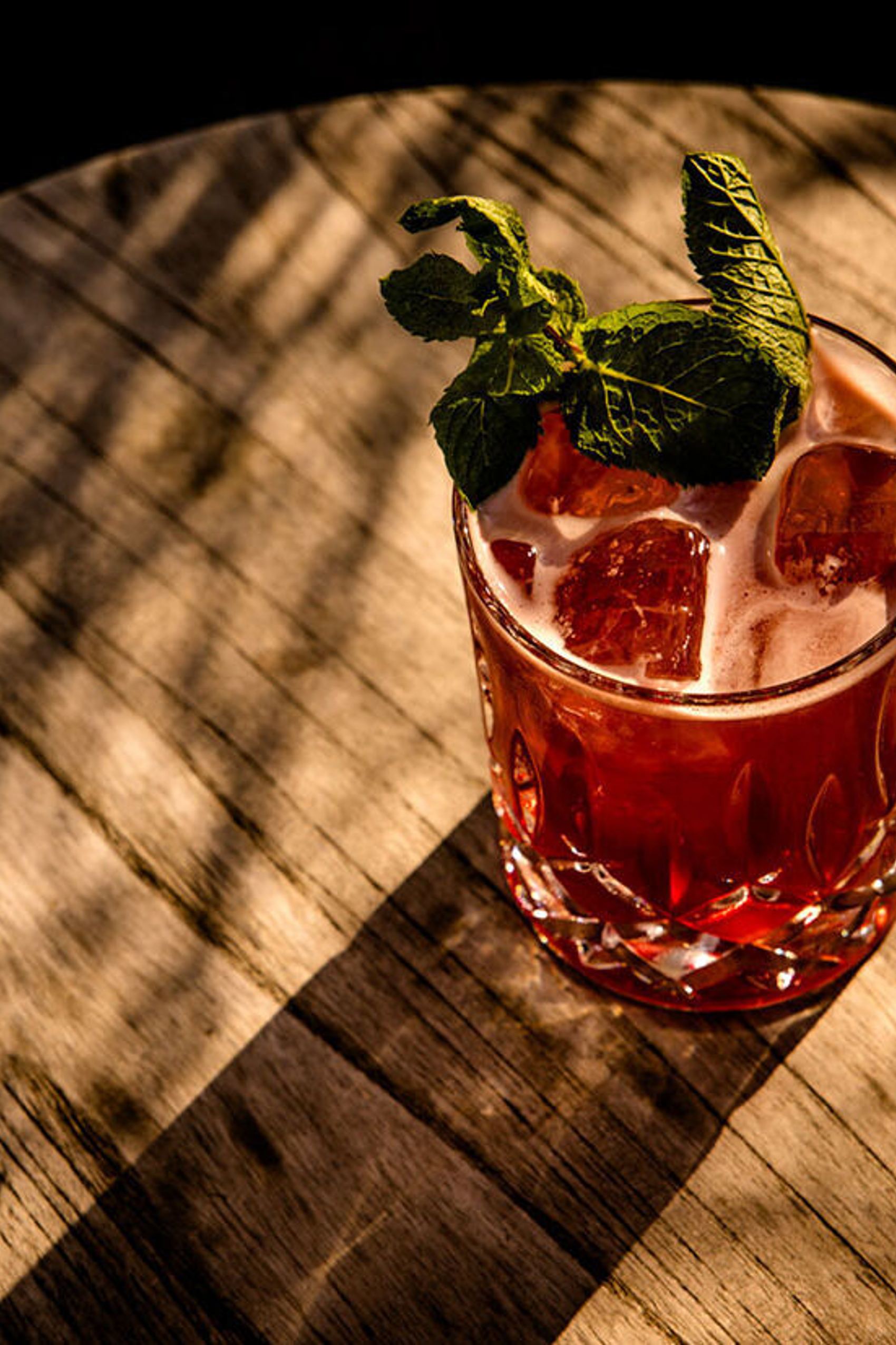 Ein Glas roter Cocktail mit Eiswürfeln und Minzblatt auf einem holzgemusterten Tisch, beleuchtet von warmem Sonnenlicht mit Schattenmustern.