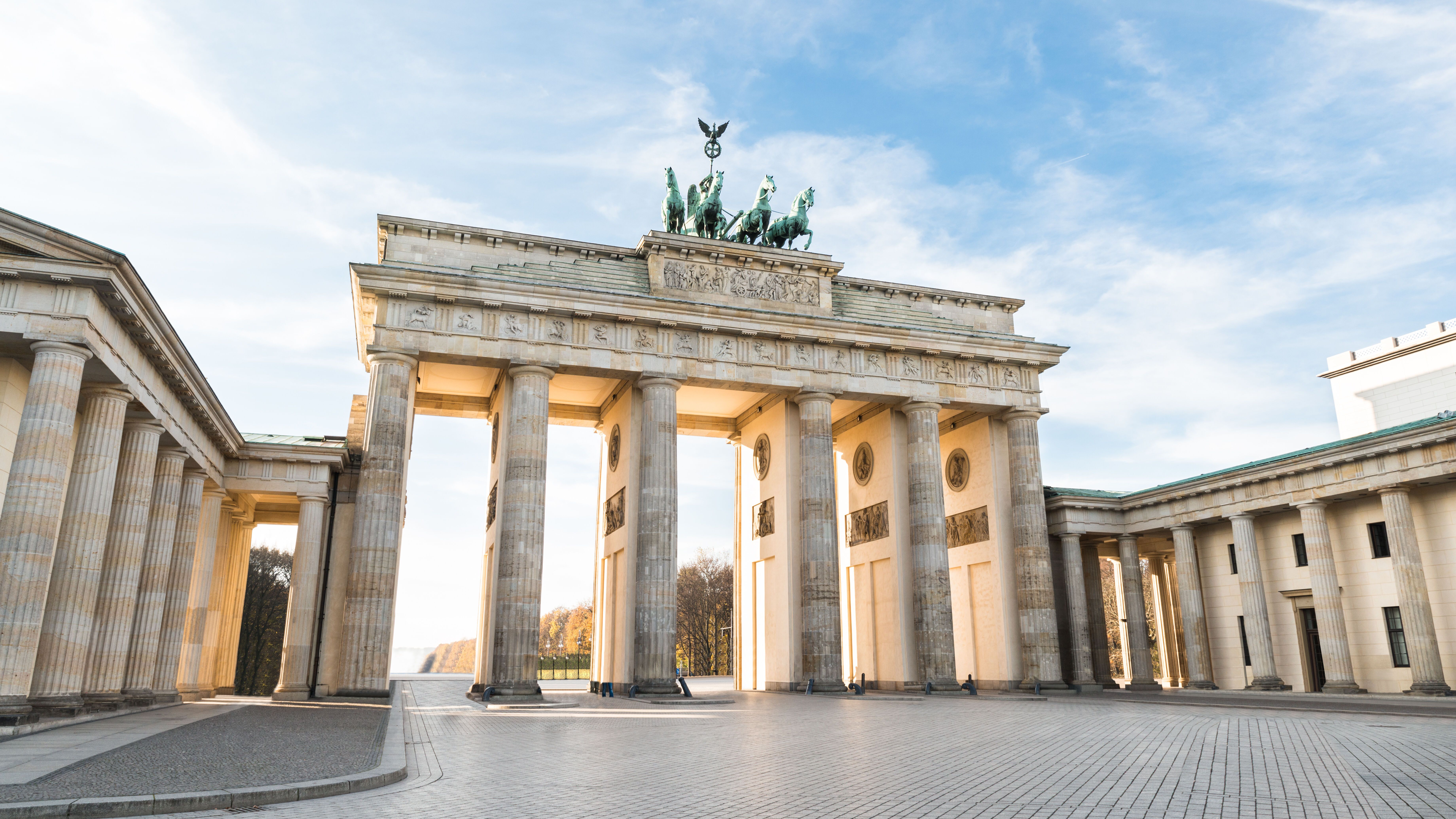 Brandenburger Tor in Berlin bei Tageslicht, mit hellen Säulen und der markanten Quadriga-Skulptur auf dem Dach. Der offene Platz vor dem Tor ist menschenleer und der Himmel zeigt leichte Wolken.