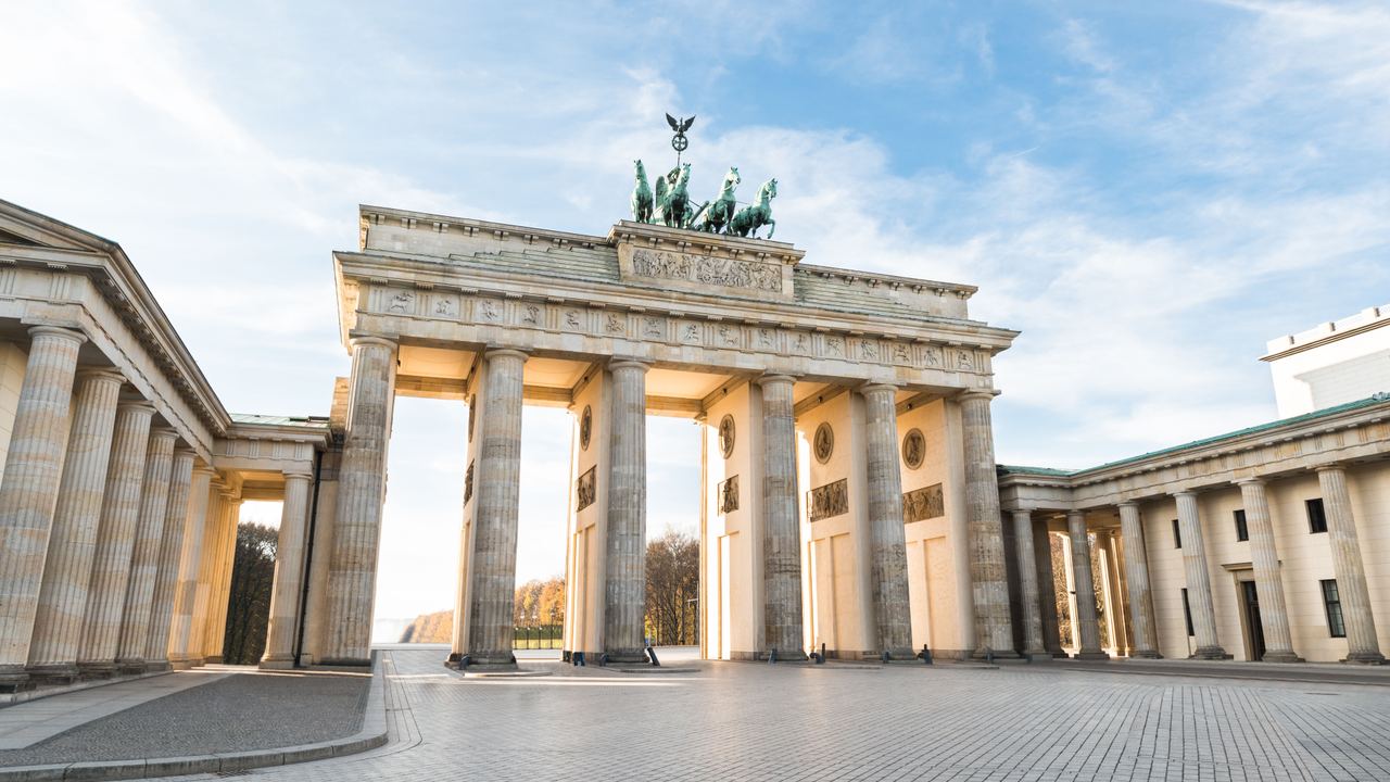 Brandenburger Tor in Berlin bei Tageslicht, mit hellen Säulen und der markanten Quadriga-Skulptur auf dem Dach. Der offene Platz vor dem Tor ist menschenleer und der Himmel zeigt leichte Wolken.