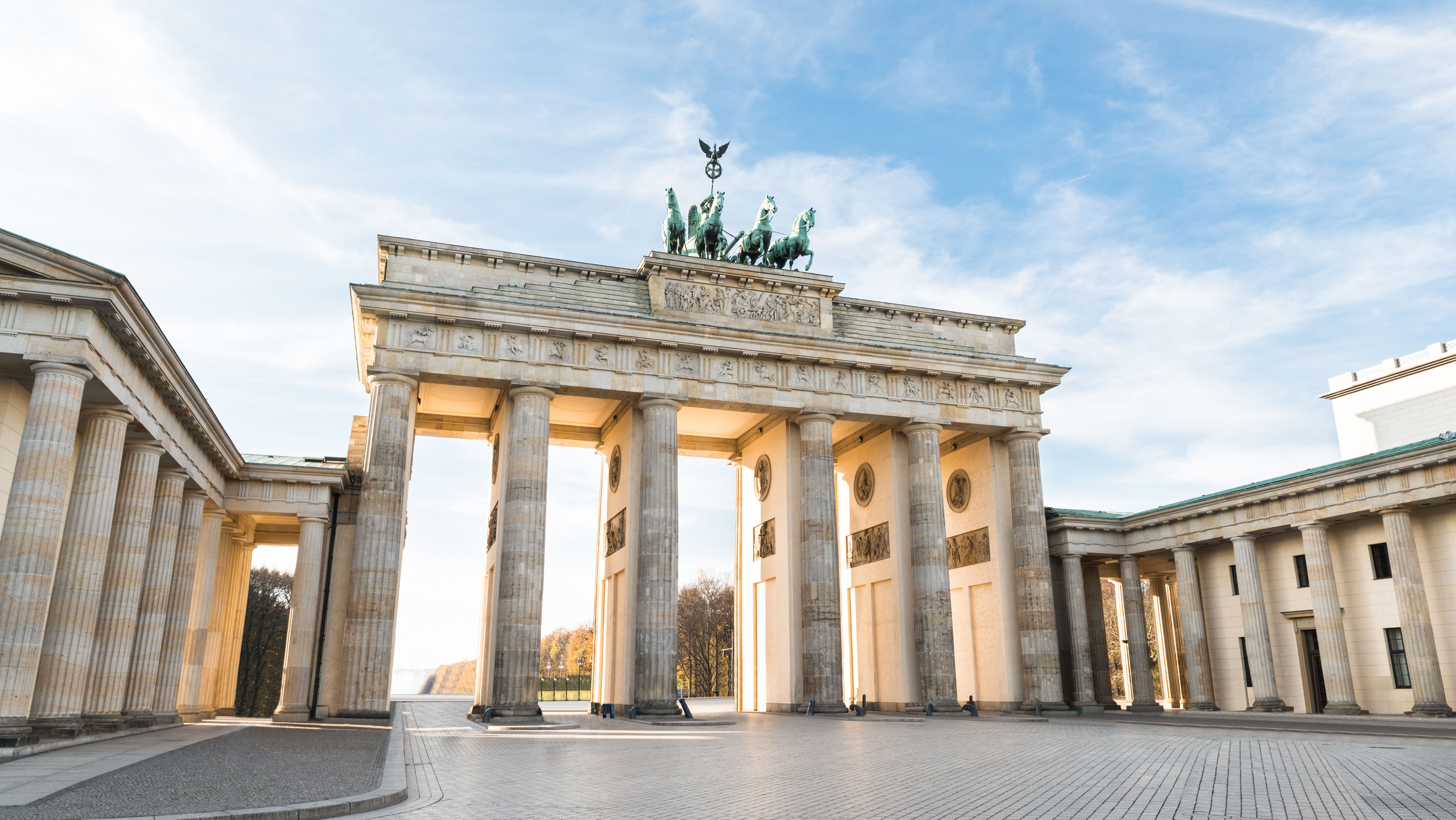 Brandenburger Tor in Berlin bei Tageslicht, mit hellen Säulen und der markanten Quadriga-Skulptur auf dem Dach. Der offene Platz vor dem Tor ist menschenleer und der Himmel zeigt leichte Wolken.