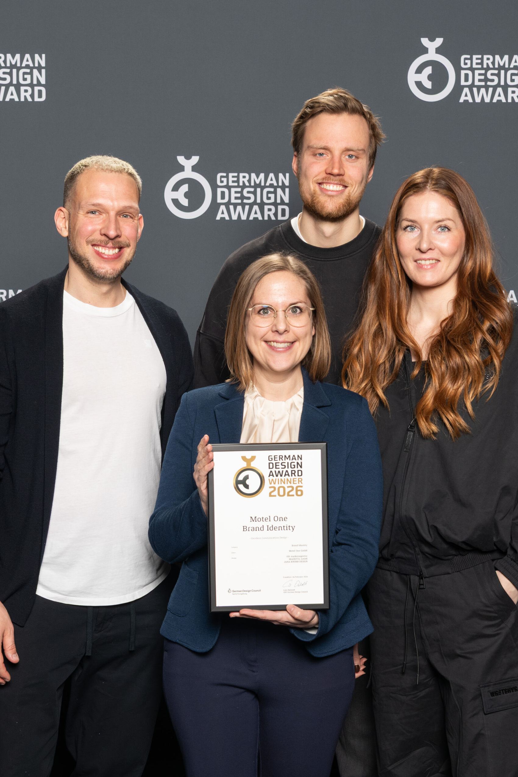 Vier Personen stehen vor einer Wand mit dem Schriftzug „German Design Award“ und halten eine Auszeichnung, die den Gewinn des German Design Awards 2026 für Motel One zeigt.