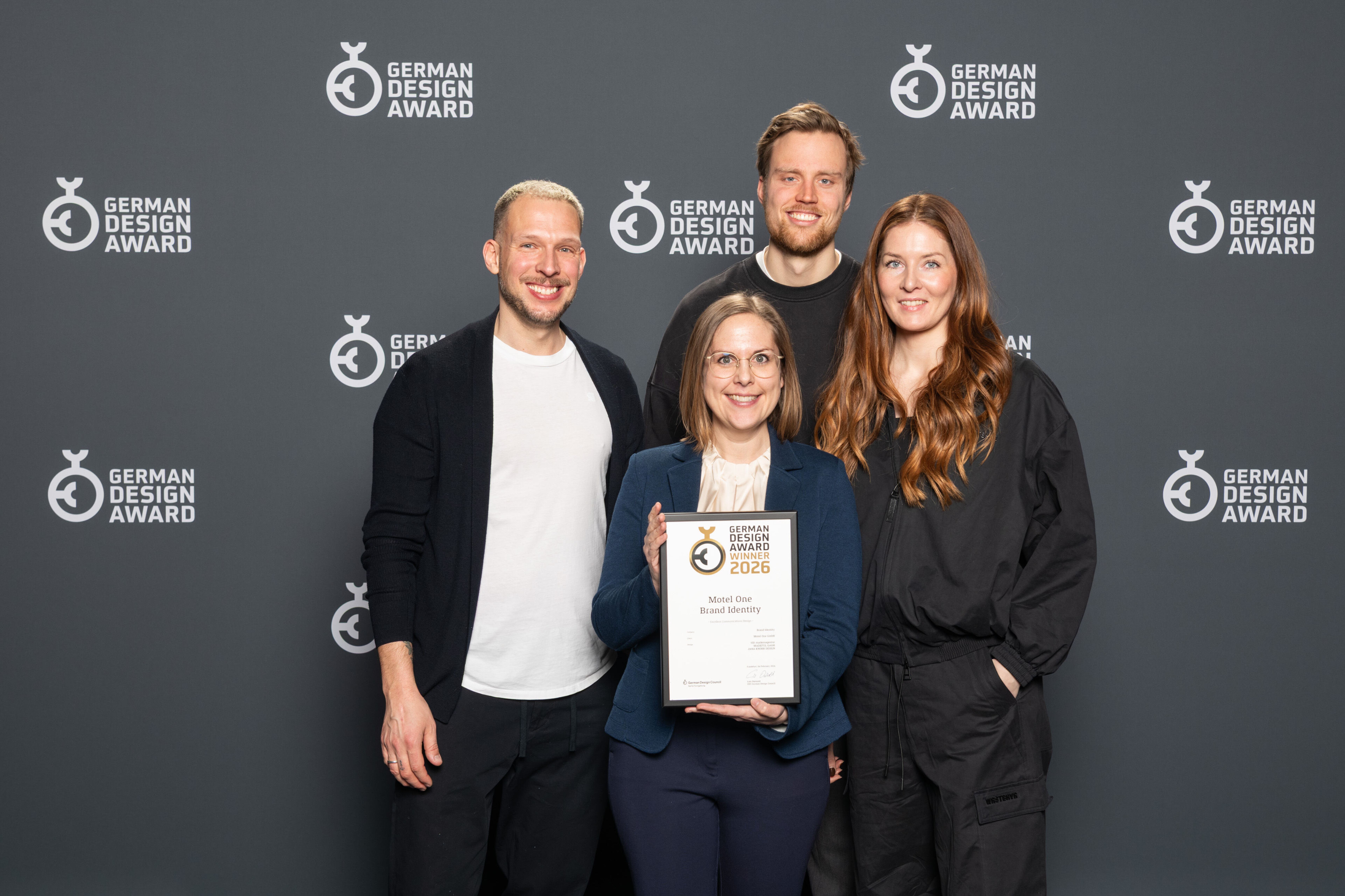 Vier Personen stehen vor einer Wand mit dem Schriftzug „German Design Award“ und halten eine Auszeichnung, die den Gewinn des German Design Awards 2026 für Motel One zeigt.