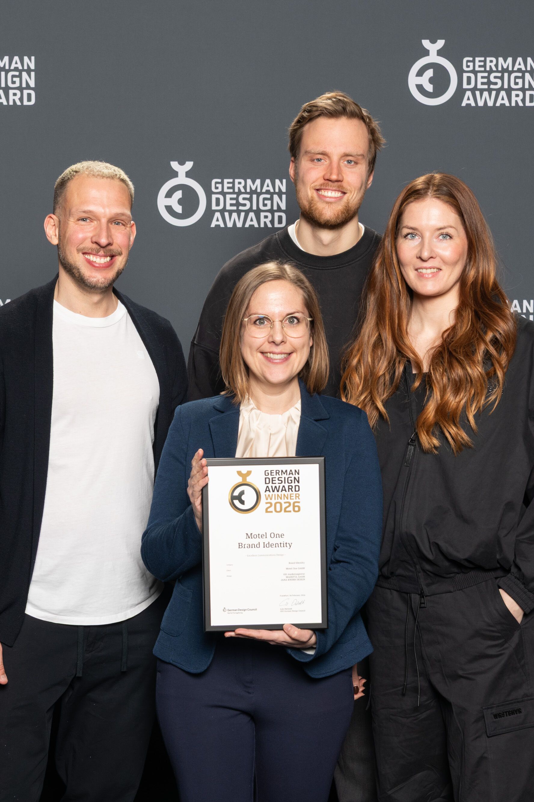 Vier Personen stehen vor einer Wand mit dem Schriftzug „German Design Award“ und halten eine Auszeichnung, die den Gewinn des German Design Awards 2026 für Motel One zeigt.