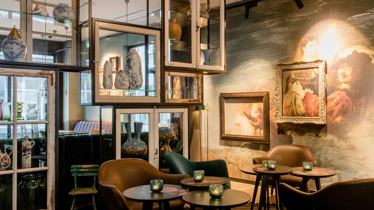 Motel One Lounge mit bequemen Sesseln und kleinen Tischen, warmes Licht, kunstvolle Wandbilder und dekorative Vasen.