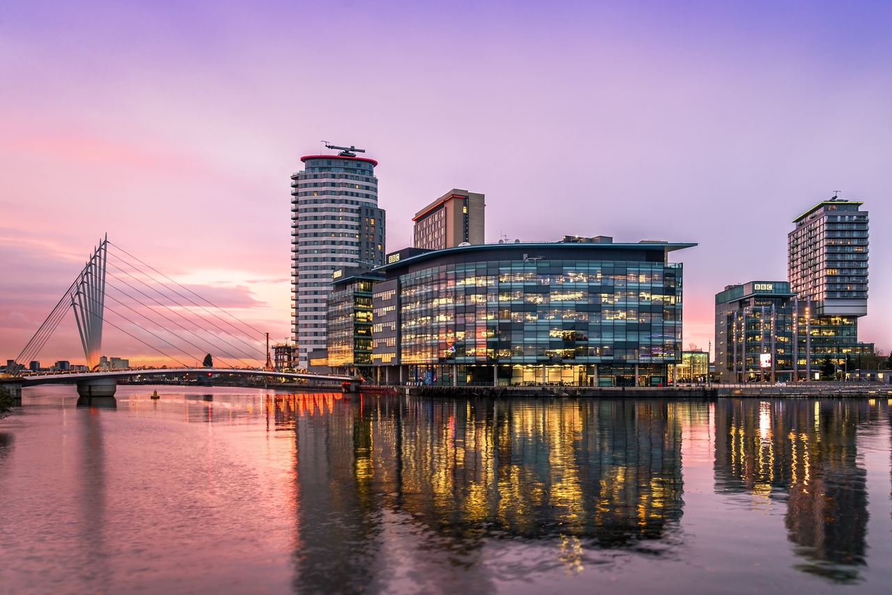 Moderne Bürogebäude am Flussufer bei Sonnenuntergang, spiegeln sich im ruhigen Wasser. Eine markante Hängebrücke verbindet die Ufer. Sanfte Abenddämmerung mit violettem und rosa Himmel schafft eine entspannte Stadtatmosphäre.