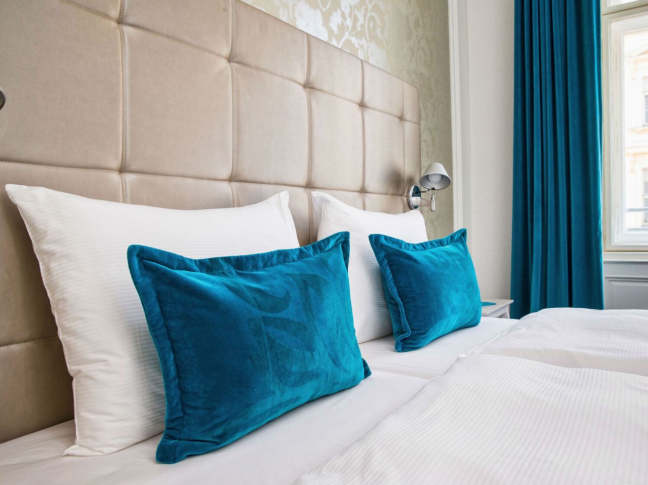 Helles, gemütliches Hotelzimmer mit großem Bett, weichen weißen Kissen und dekorativen türkisblauen Kissen. Elegantes, gepolstertes Kopfteil und Tageslicht durch Fenster mit blauen Vorhängen
