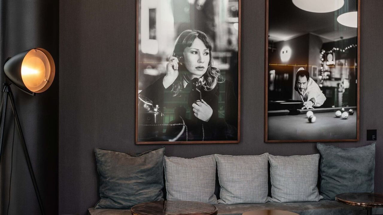 Motel One Hotel-Loungebereich mit bequemer Sitzbank, Kissen und stilvoller Stehlampe. An der Wand hängen zwei großformatige Schwarz-Weiß-Porträts, die eine entspannte und elegante Atmosphäre schaffen.