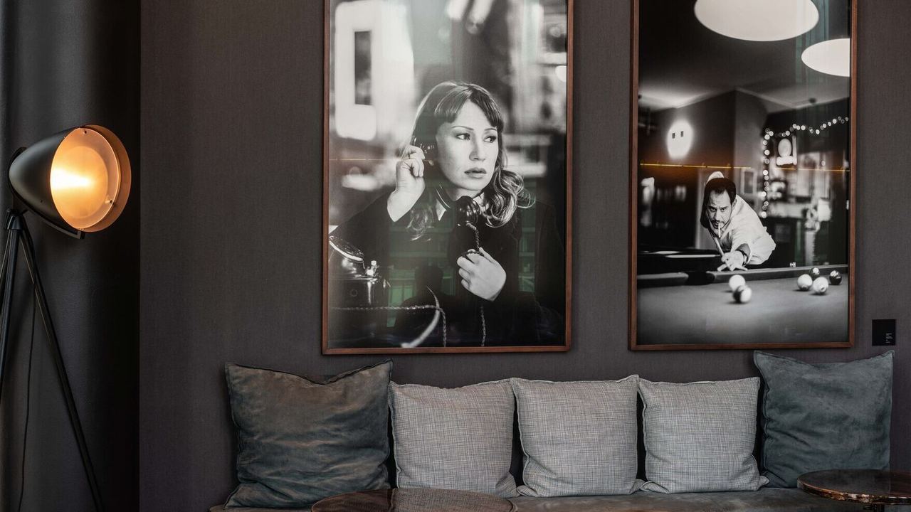 Motel One Hotel-Loungebereich mit bequemer Sitzbank, Kissen und stilvoller Stehlampe. An der Wand hängen zwei großformatige Schwarz-Weiß-Porträts, die eine entspannte und elegante Atmosphäre schaffen.
