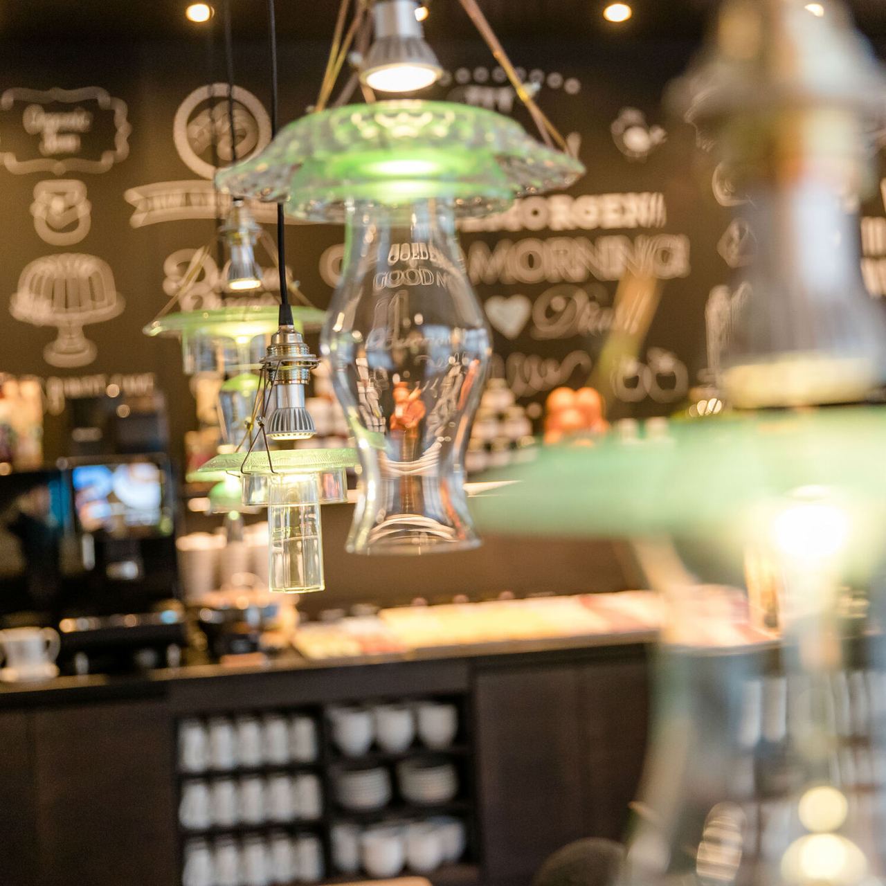 Nahaufnahme stilvoller, grüner Hängelampen in einem gemütlichen Café- oder Hotelrestaurant mit warmer, einladender Atmosphäre und dekorativ gestalteter Wand im Hintergrund.