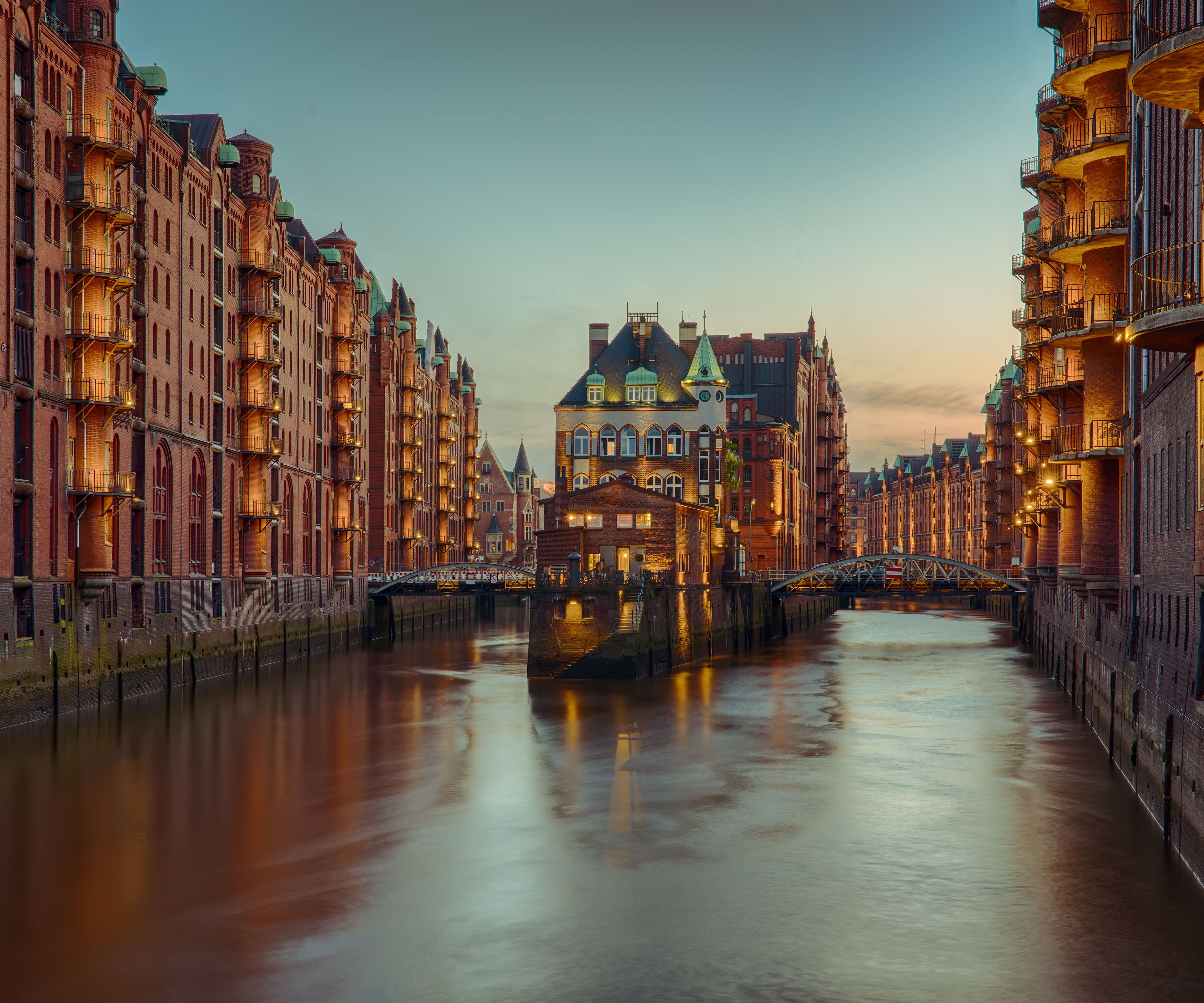 Blick auf einen ruhigen Kanal umgeben von historischen Backsteingebäuden bei Sonnenuntergang, mit warmem Licht, das die Fassaden erhellt und eine einladende, friedliche Atmosphäre schafft.