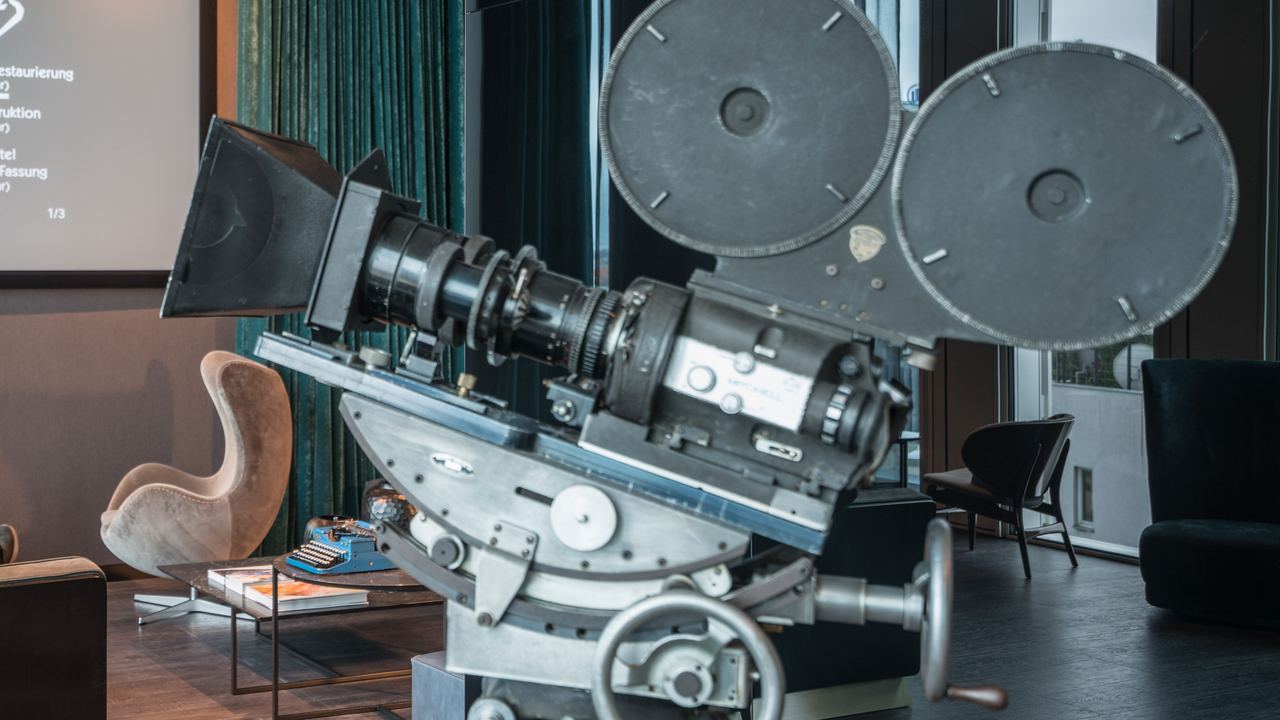 Designhotel Berlin Upper West Motel One Lounge mit zwei Personen, die mit großen Kopfhörern auf einem Sofa sitzen und auf eine Leinwand mit Film schauen sowie eine Filmkamera im Vordergrund